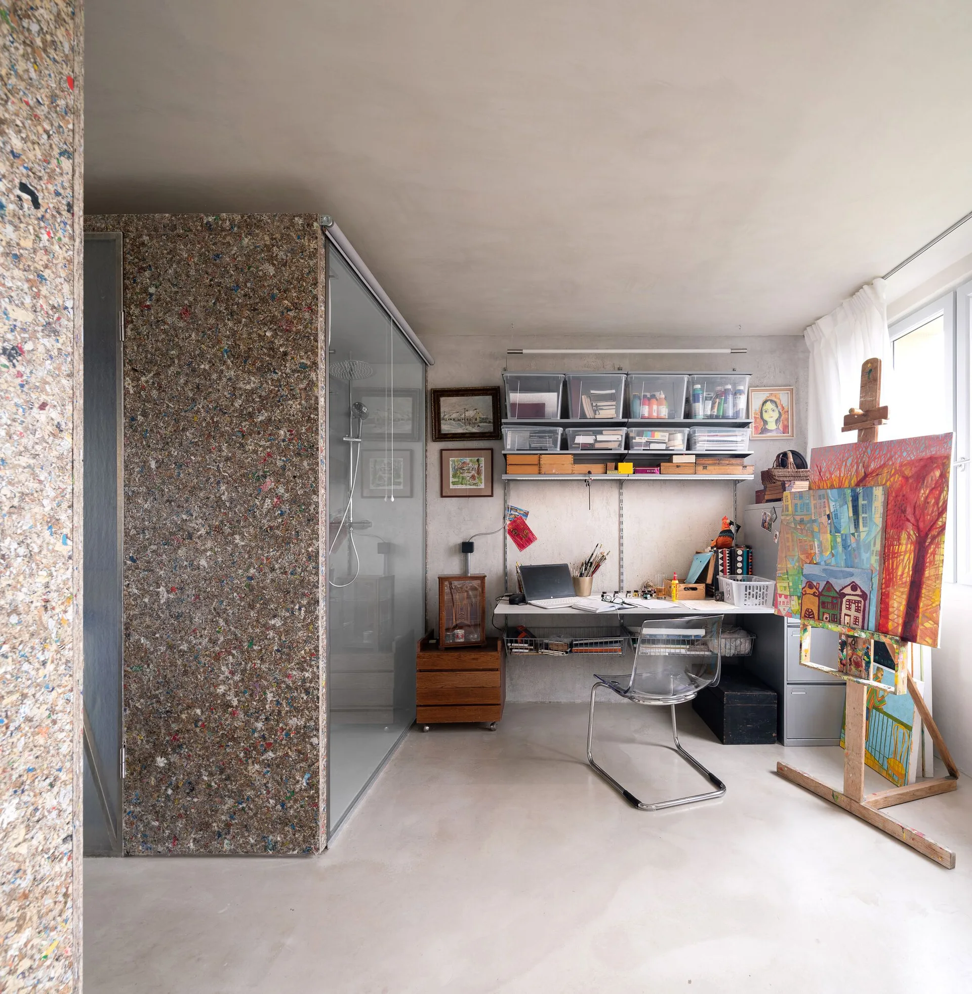 A creative home workspace with a wall-mounted desk, a clear plastic chair, an art easel with a painting, and a speckled recycled composite partition.