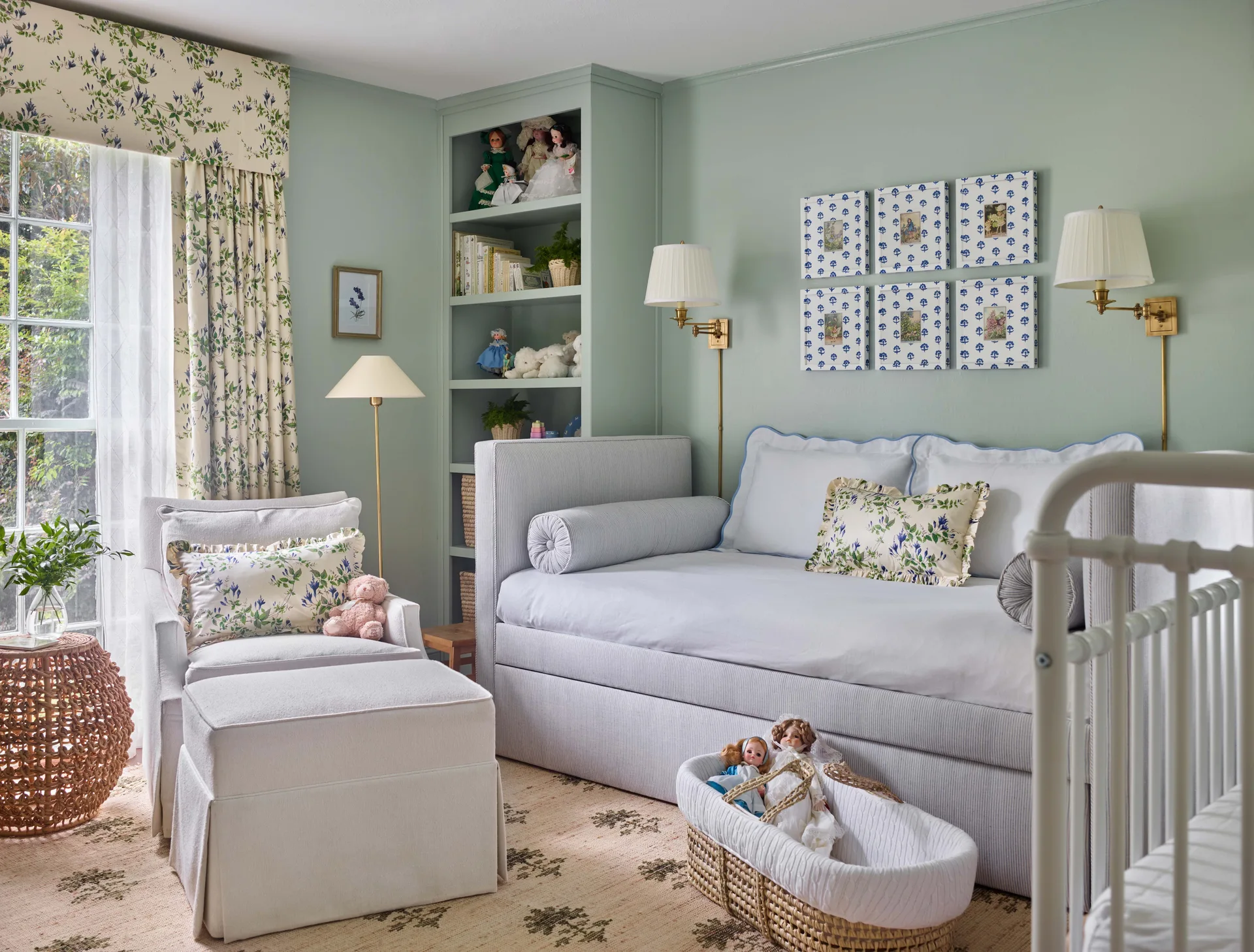 A traditional sage green nursery features a striped upholstered daybed, an armchair with an ottoman, a metal crib, and floral drapes.
