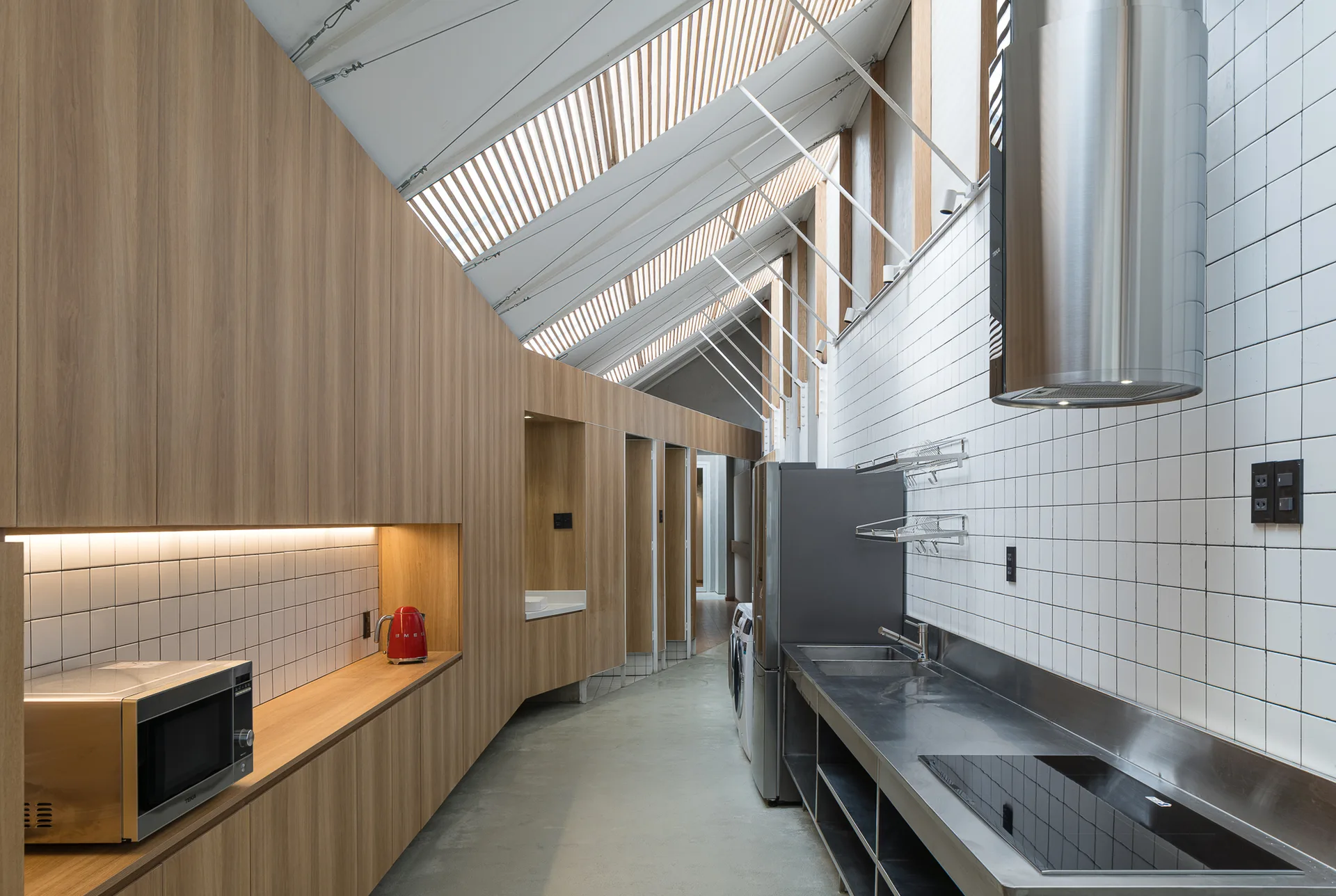 A modern galley kitchen featuring a slanted wood-slat skylight ceiling, long wood cabinets, stainless steel countertops, and a white tiled wall.