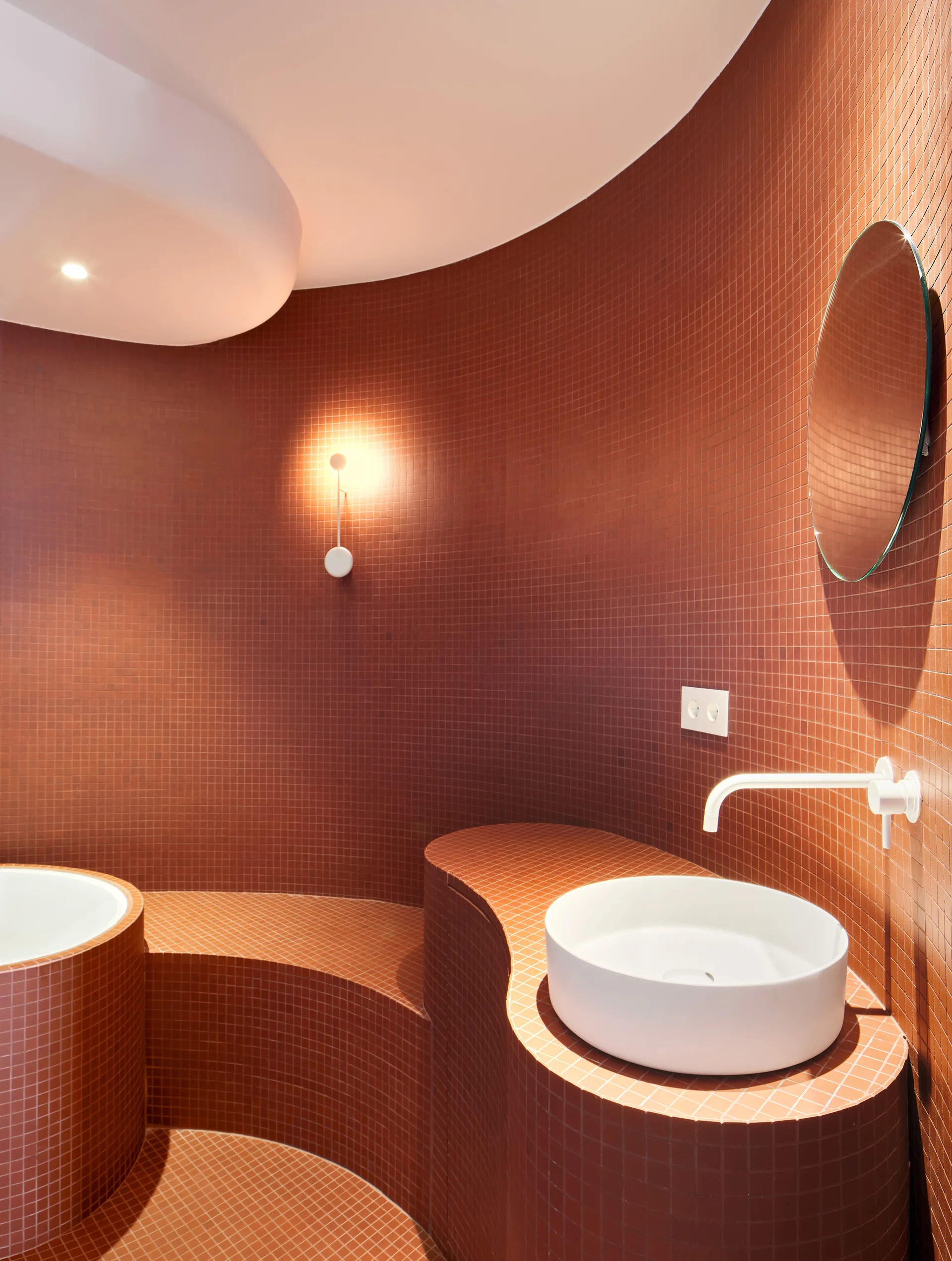 A bathroom with curved walls and surfaces clad in red terracotta mosaic tiles features a white vessel sink, a round mirror, and a wall sconce.