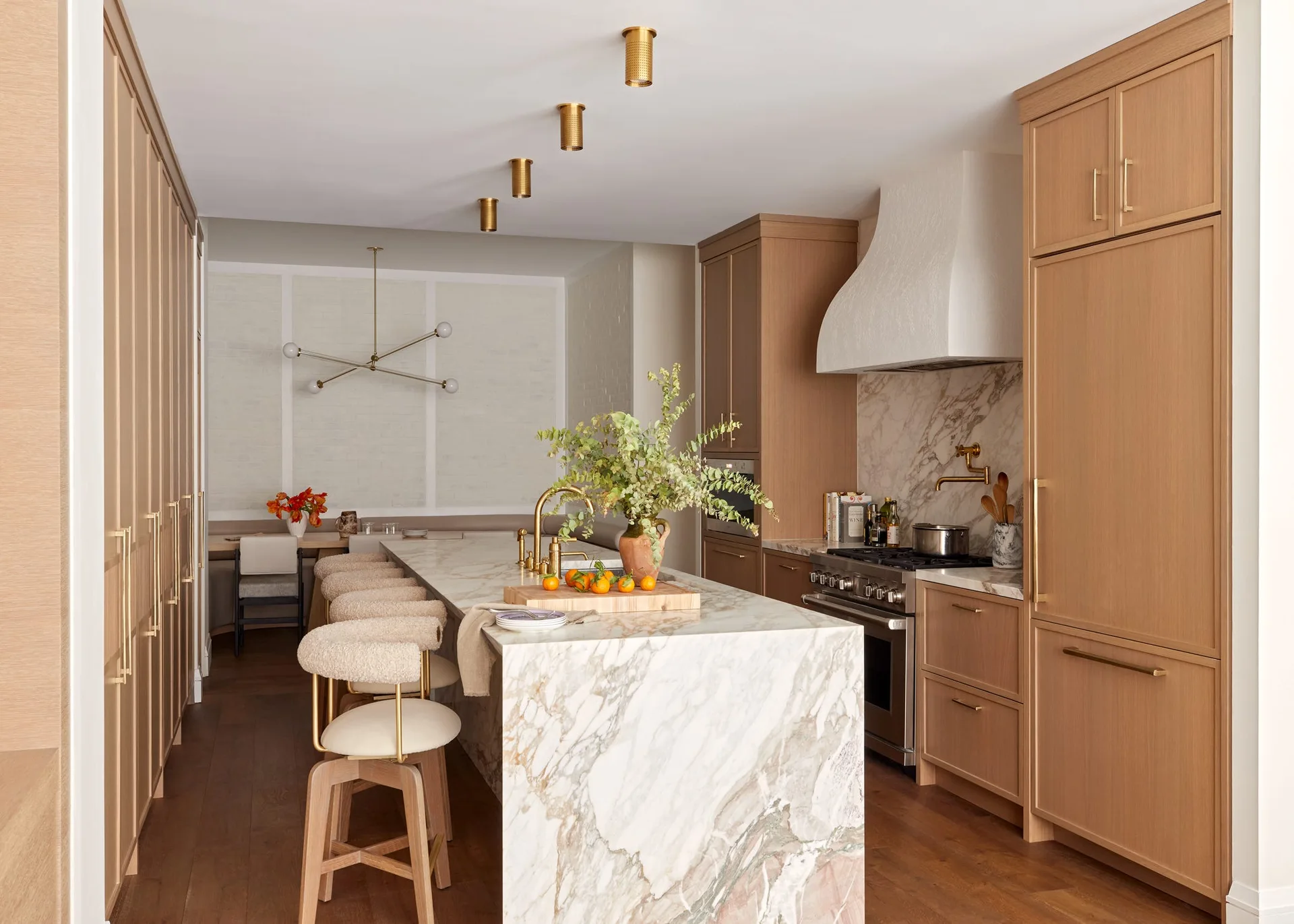 A kitchen featuring a marble-topped waterfall island with bouclé stools, flat-panel oak cabinetry, and brass ceiling light fixtures.