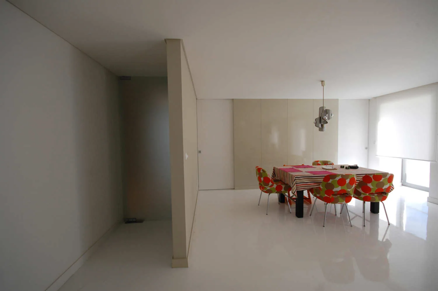 A minimalist dining room features a table with patterned polka-dot chairs, a glossy white floor, a beige partition wall, and built-in cabinetry.