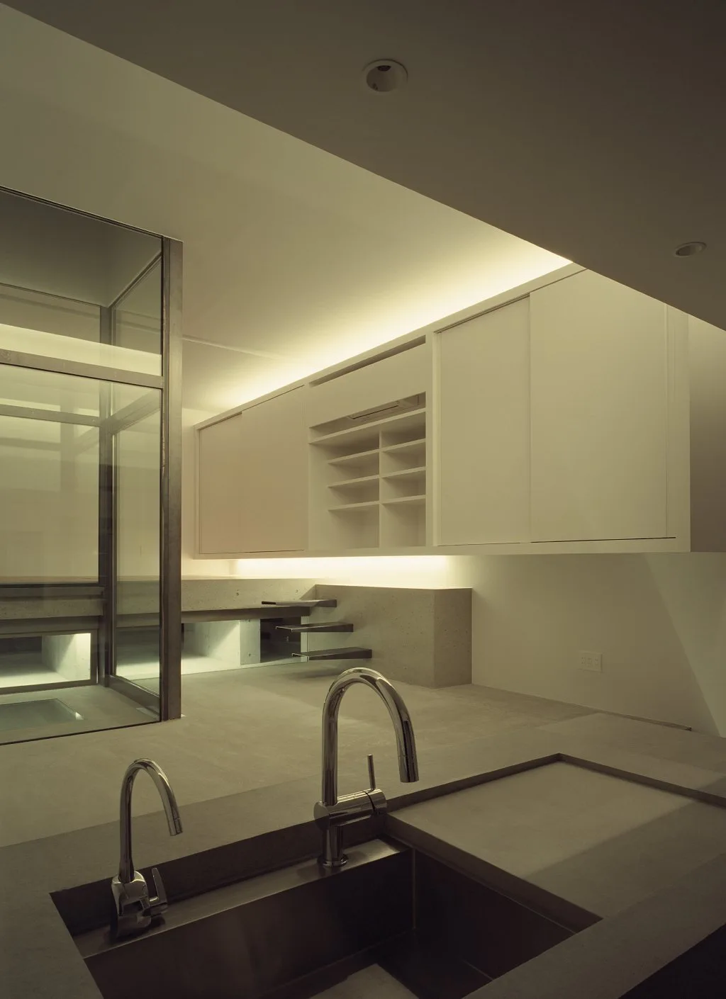 A minimalist grey kitchen with a concrete countertop, dual chrome faucets, white floating cabinets, and integrated warm cove lighting.