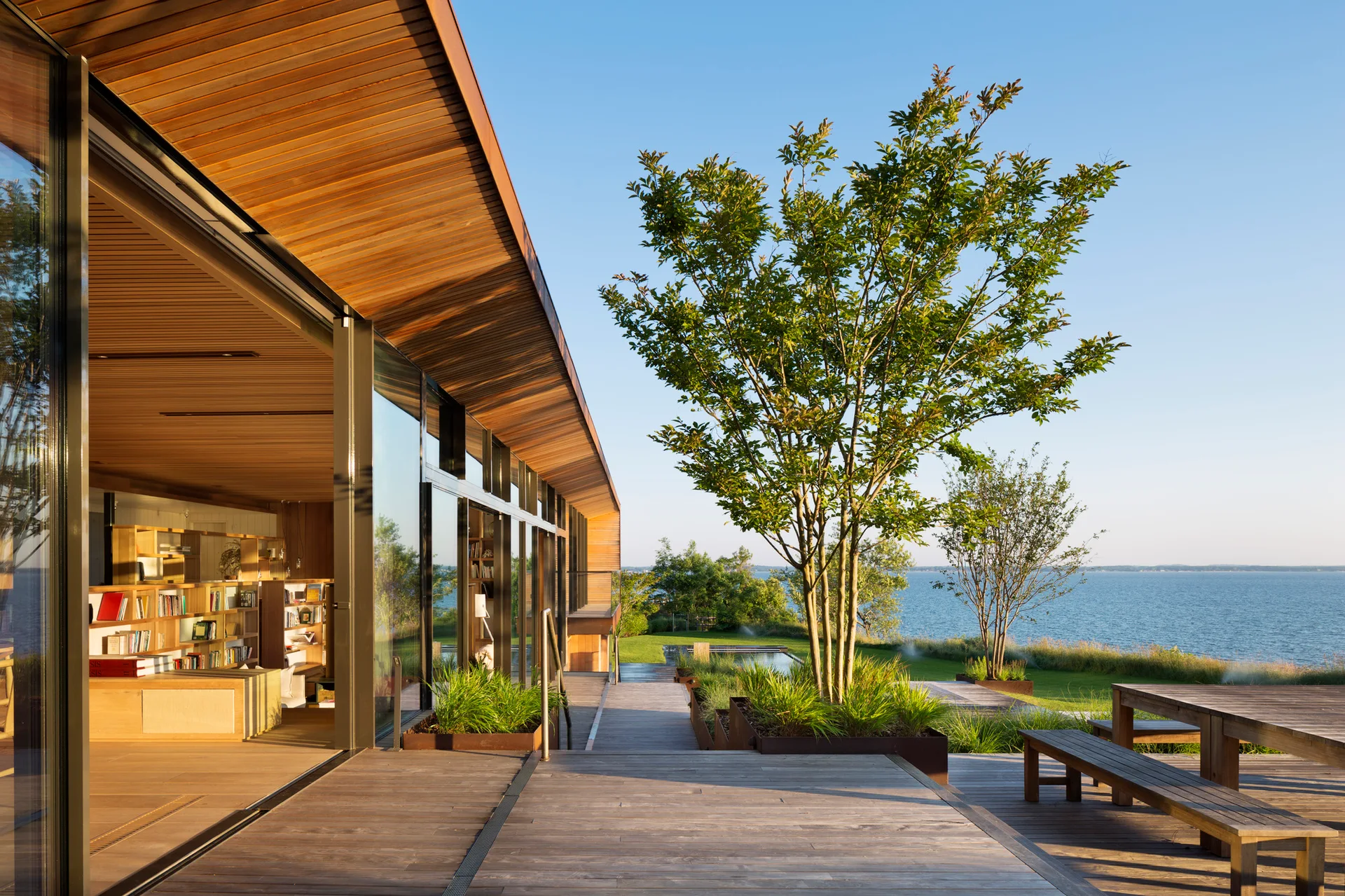 A modern waterfront home features a wood plank deck, sliding glass doors, integrated metal planters with trees, and a slatted timber ceiling overhang.