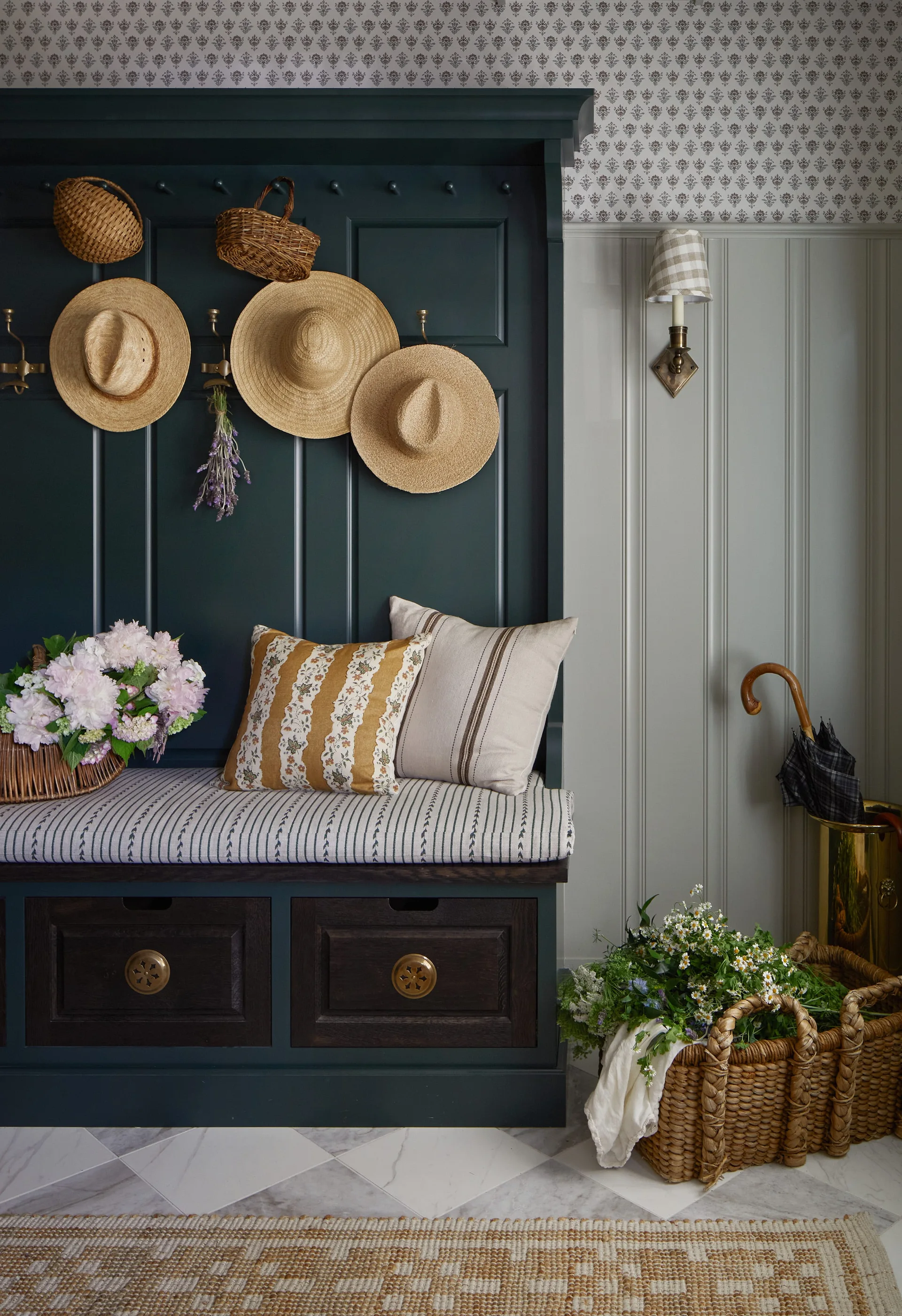 A dark green built-in entryway bench with a striped cushion is adorned with hanging straw hats, decorative pillows, and a flower basket.