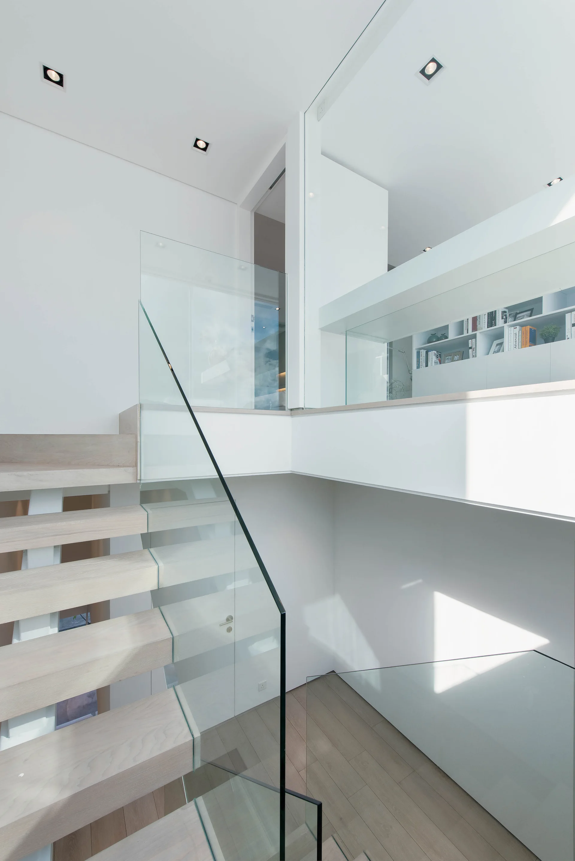 A modern floating staircase with light wood treads and a clear glass railing rises up to a white mezzanine landing with built-in bookshelves.