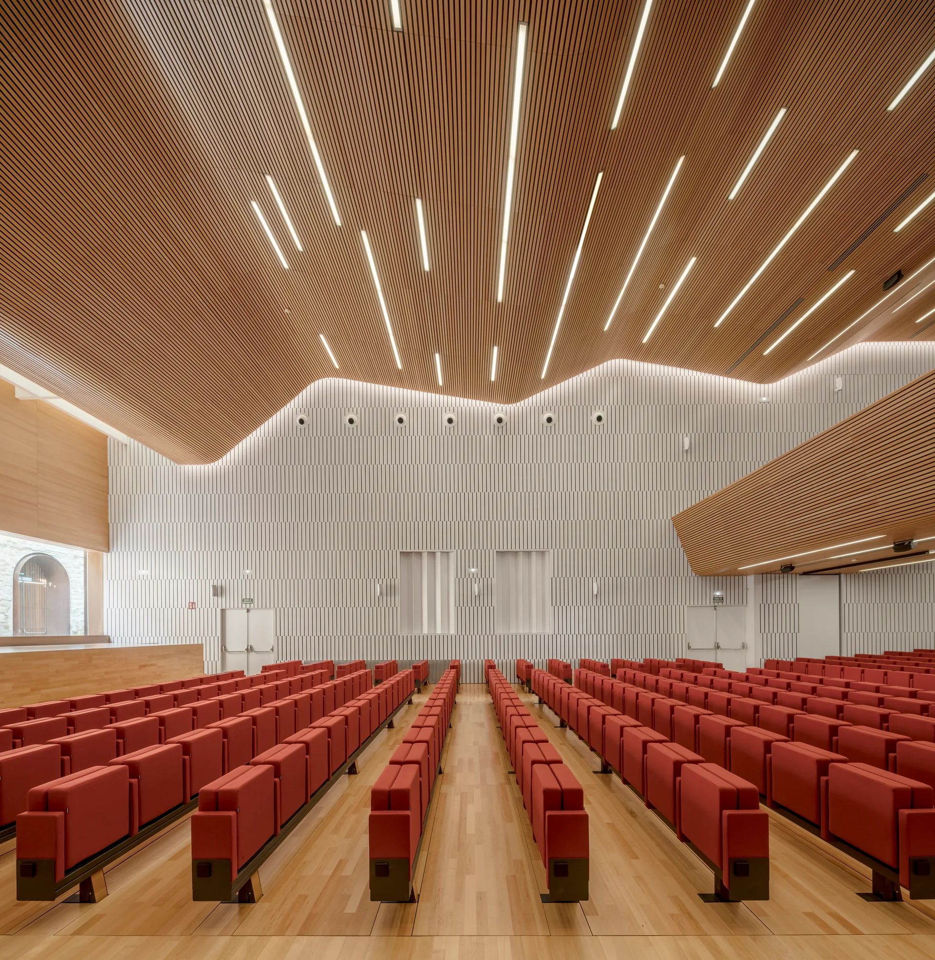 An empty auditorium features rows of red seating beneath an undulating wood-slatted ceiling with built-in linear light strips and white textured walls.