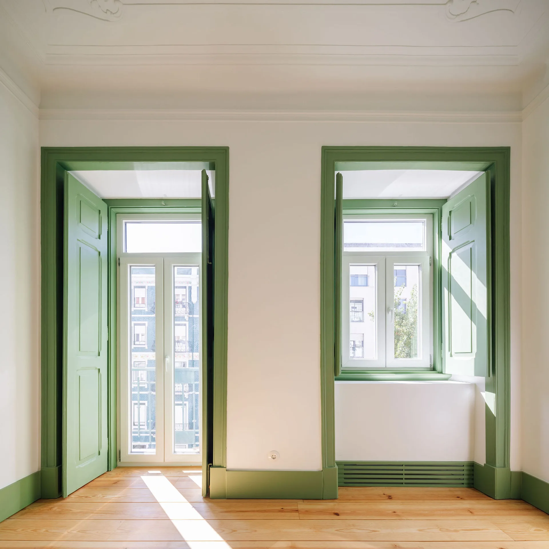 Two large windows with sage green trim and interior shutters set against plain white walls and natural light wood flooring.