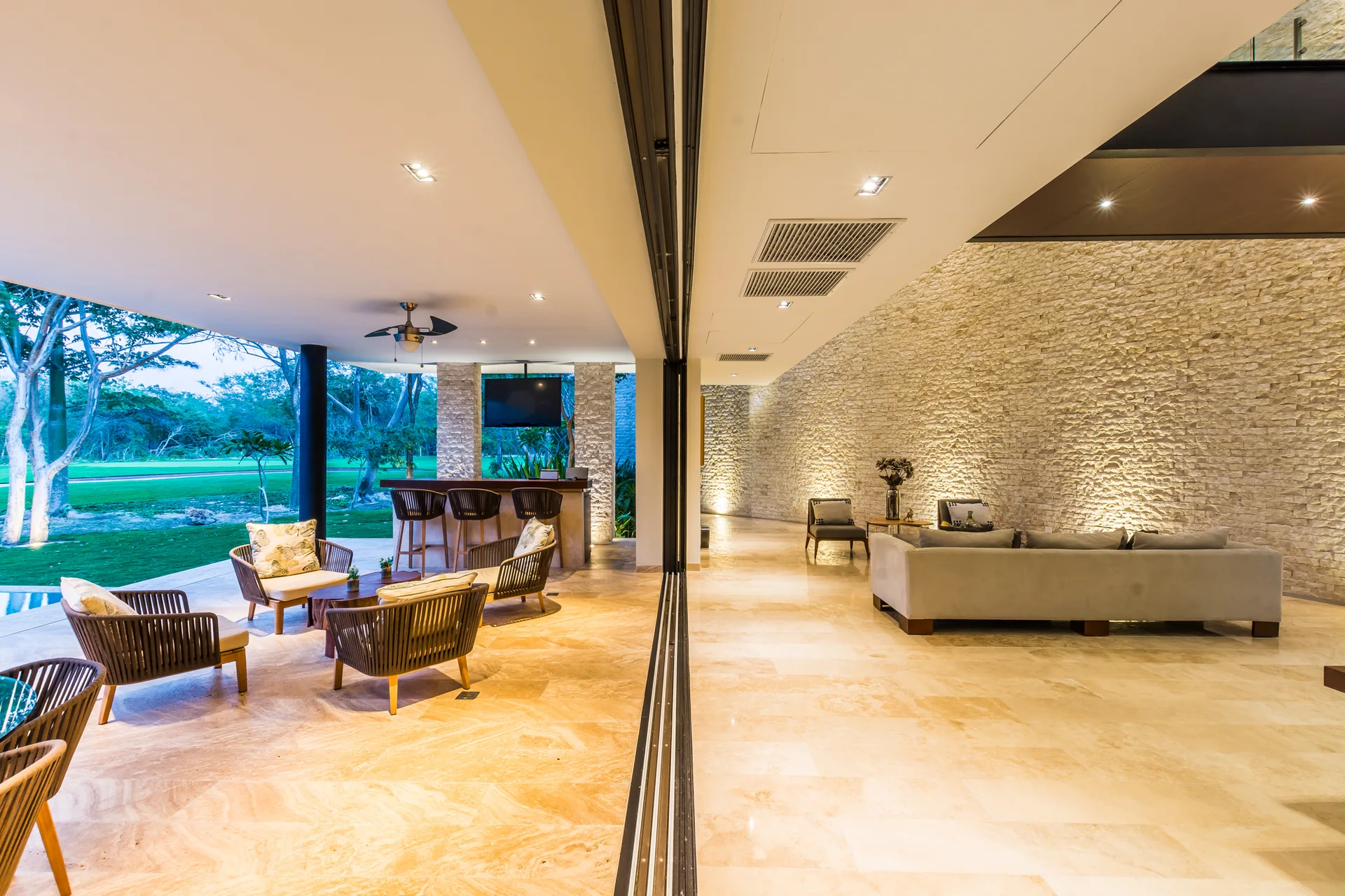 An open-plan living room with a grey sofa and a textured stone wall connects seamlessly to an outdoor patio with wicker seating.