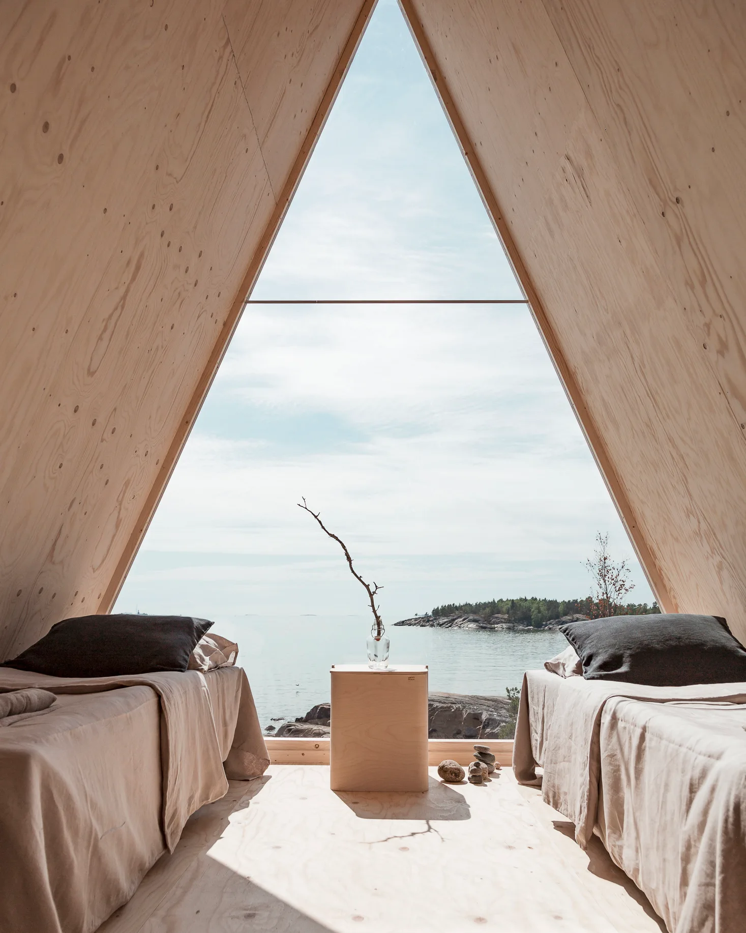 A minimalist A-frame cabin bedroom features two twin beds with grey pillows, a central wooden side table, and a large triangular window overlooking the sea.