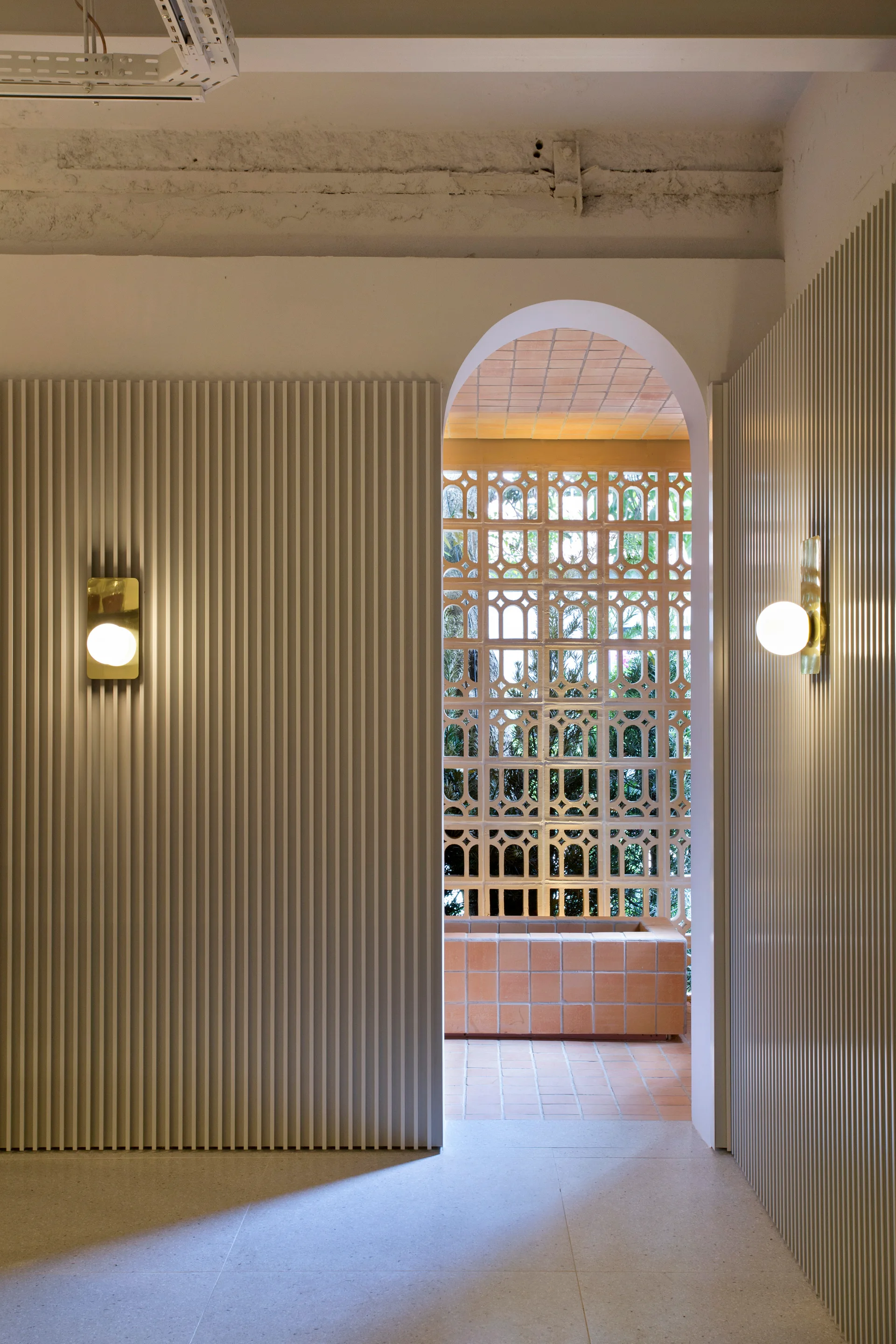 White fluted wall panels with globe sconces flank an arched opening that looks into a terracotta tiled courtyard with a decorative breeze block wall.