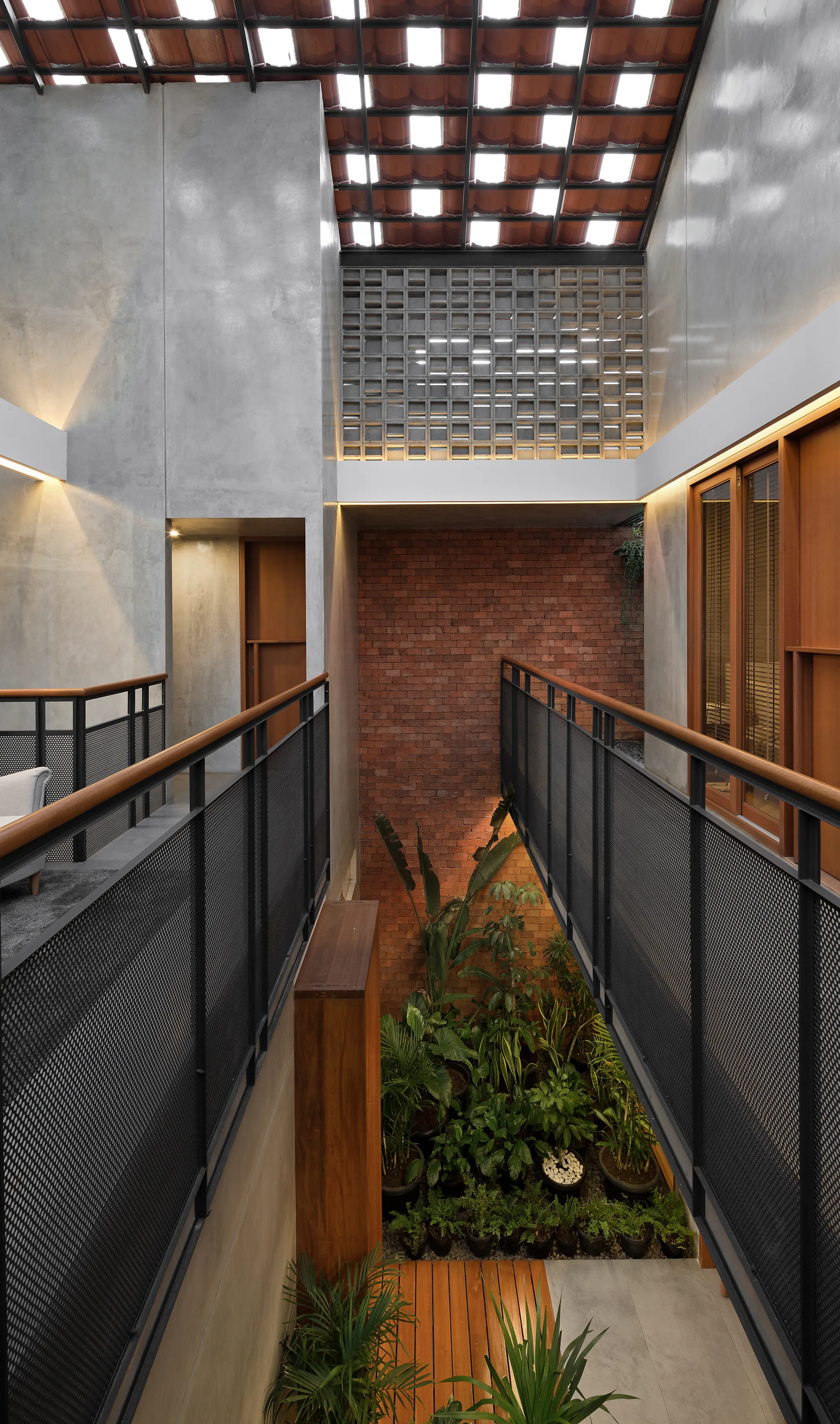 An elevated black bridge walkway overlooks a sunken indoor garden with potted plants, an exposed brick wall, and smooth concrete surfaces.