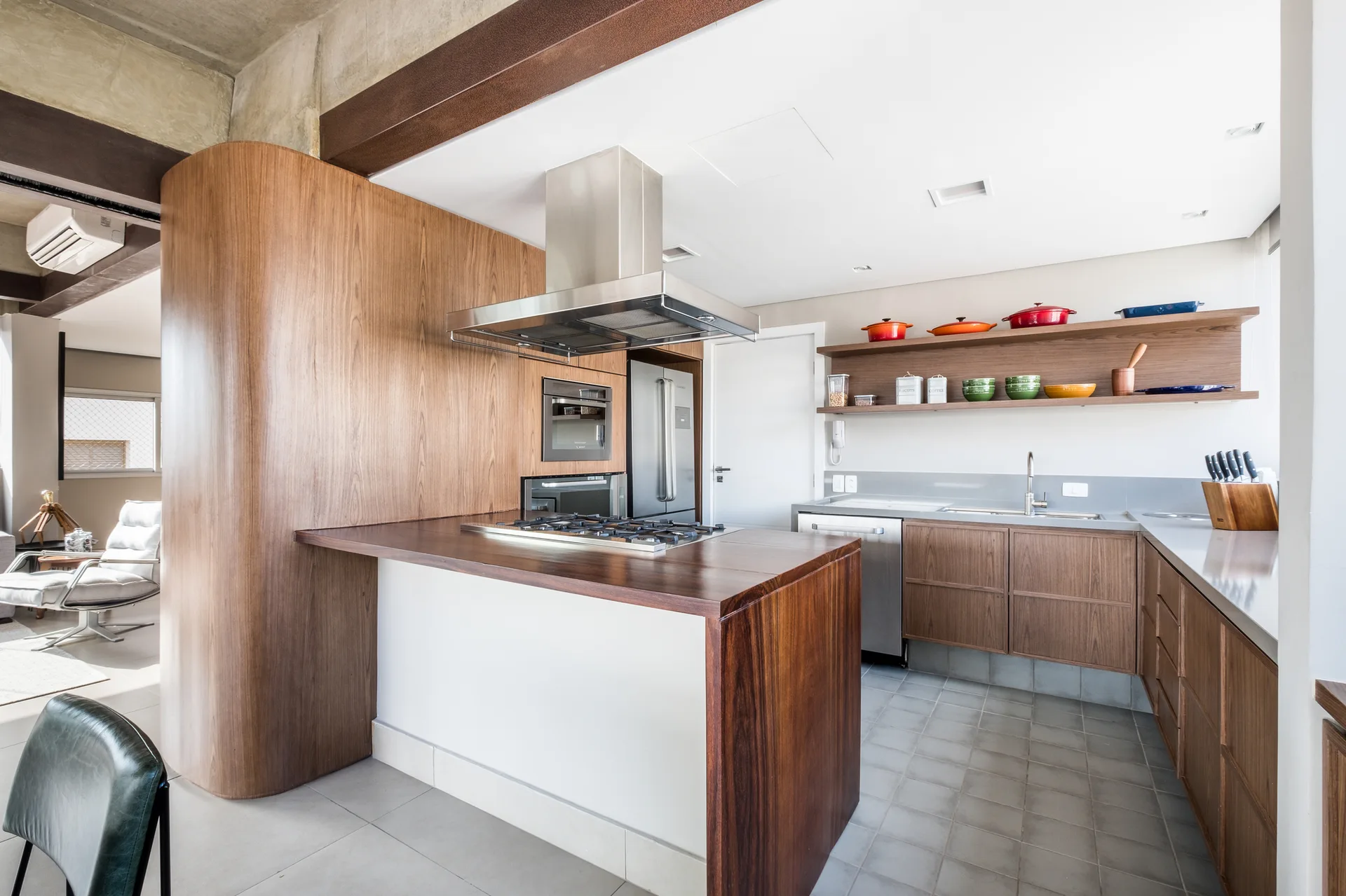 A modern kitchen featuring a wood-topped island with a cooktop, curved wood wall paneling, open shelves with colorful cookware, and grey tiled floors.