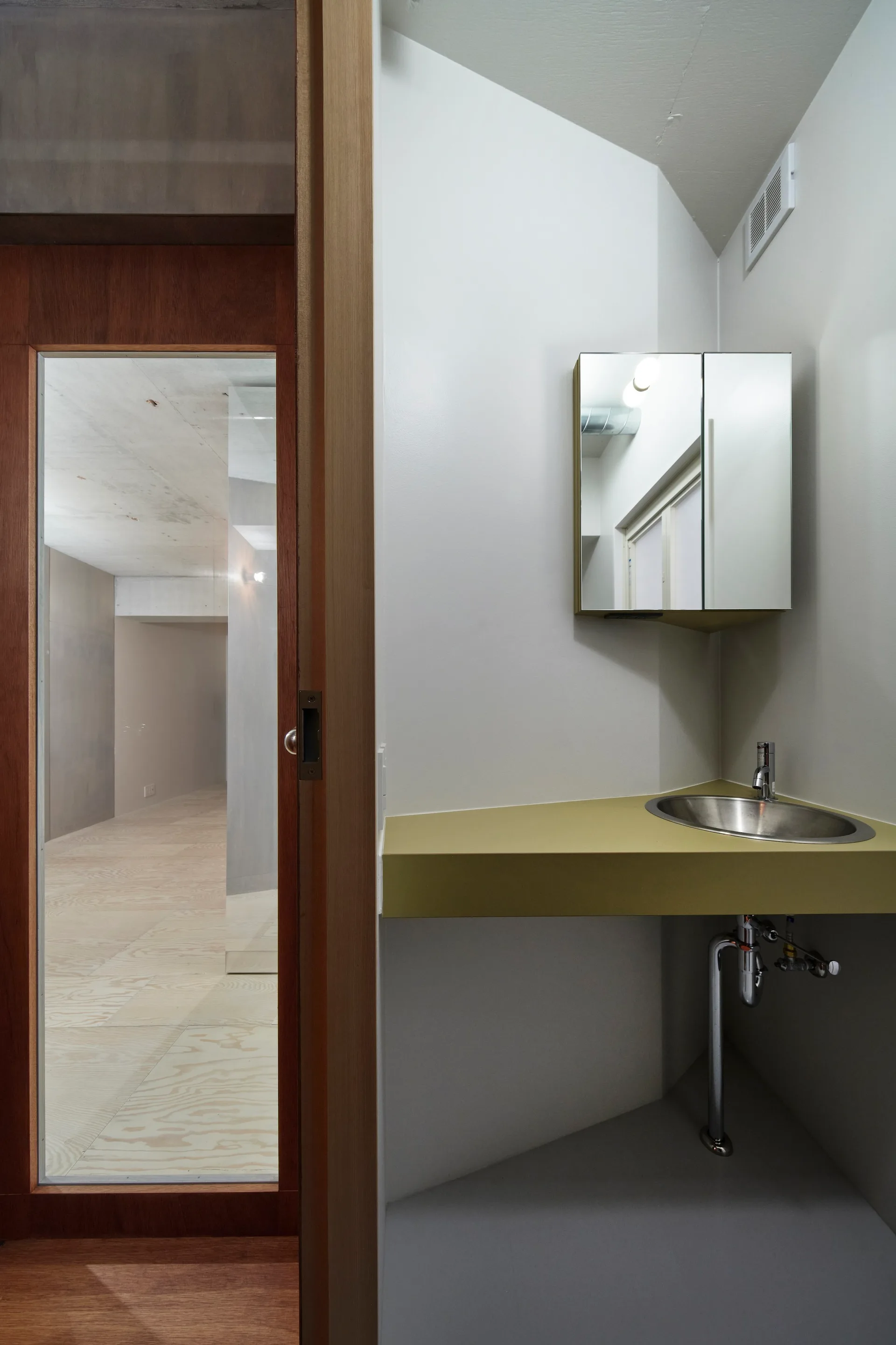A minimalist powder room features a triangular olive green corner vanity with a stainless steel sink, a mirrored cabinet, and a wooden sliding door.