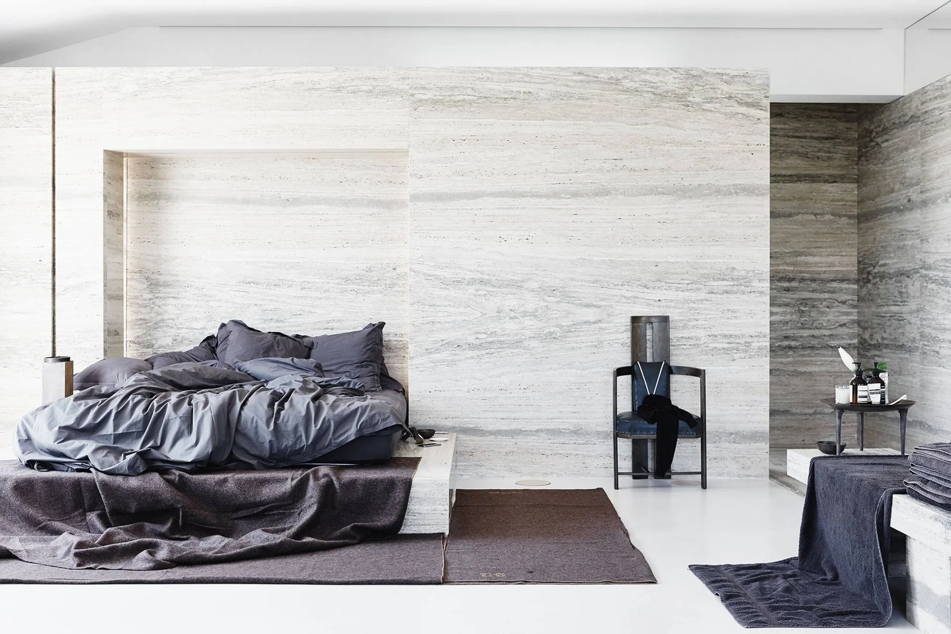 A minimalist bedroom features a low platform bed set into a recessed alcove of a large travertine stone wall.