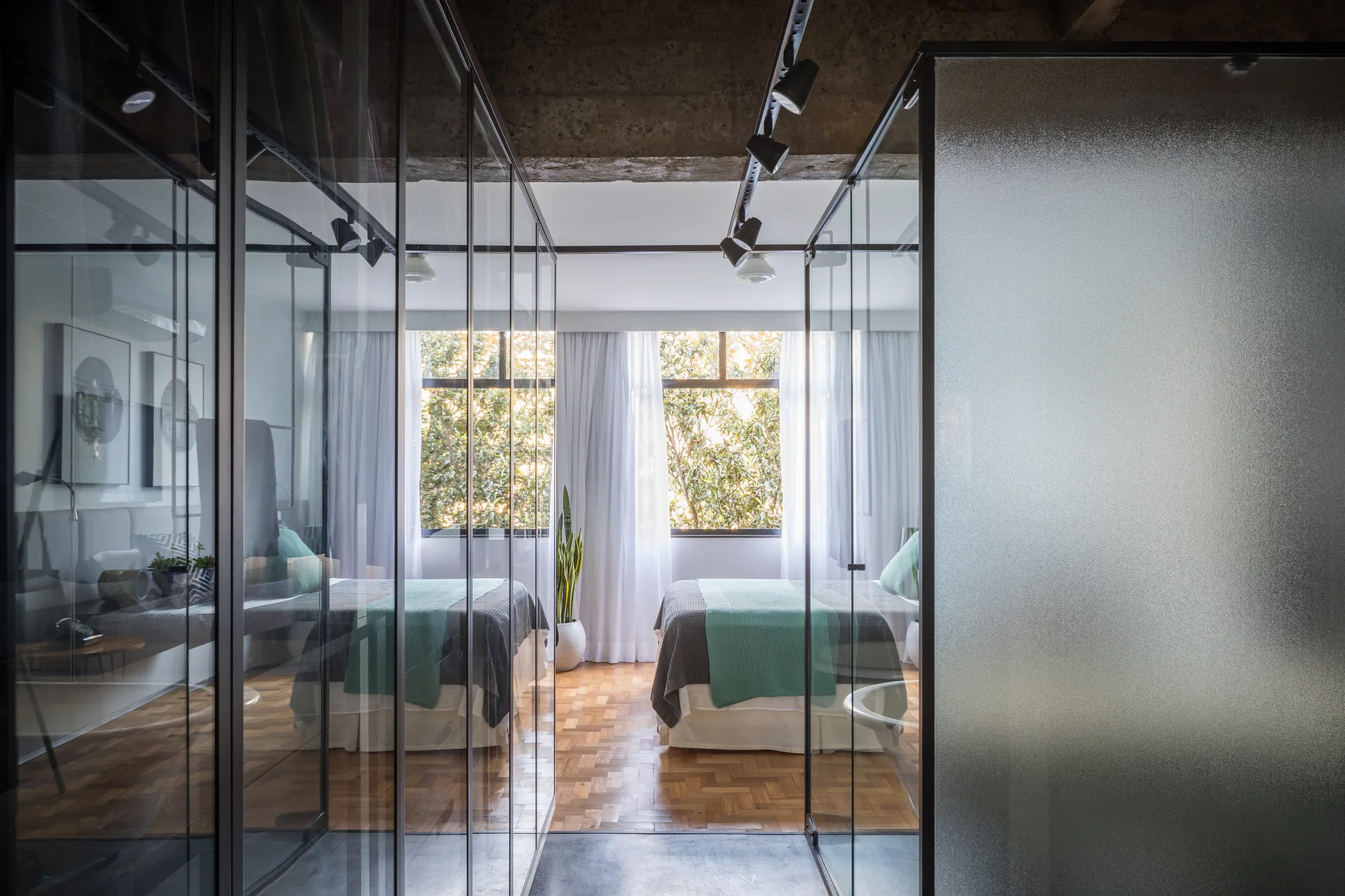 A modern bedroom viewed through glass wardrobe enclosures with two beds, wood parquet flooring, and an exposed concrete ceiling beam.