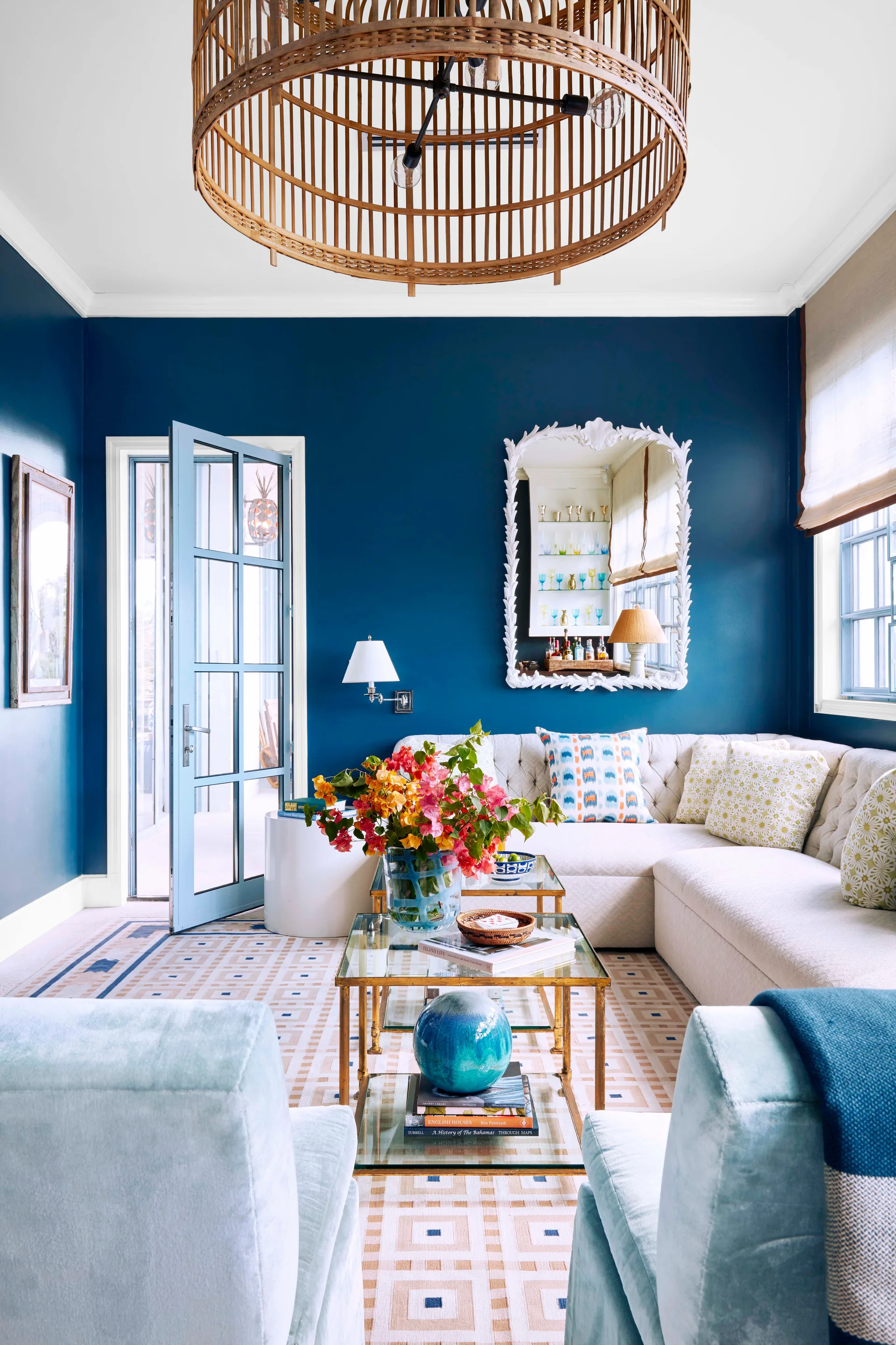 A vibrant living room featuring navy blue walls, a white tufted sectional sofa, glass-top coffee tables, and a large woven rattan pendant light.