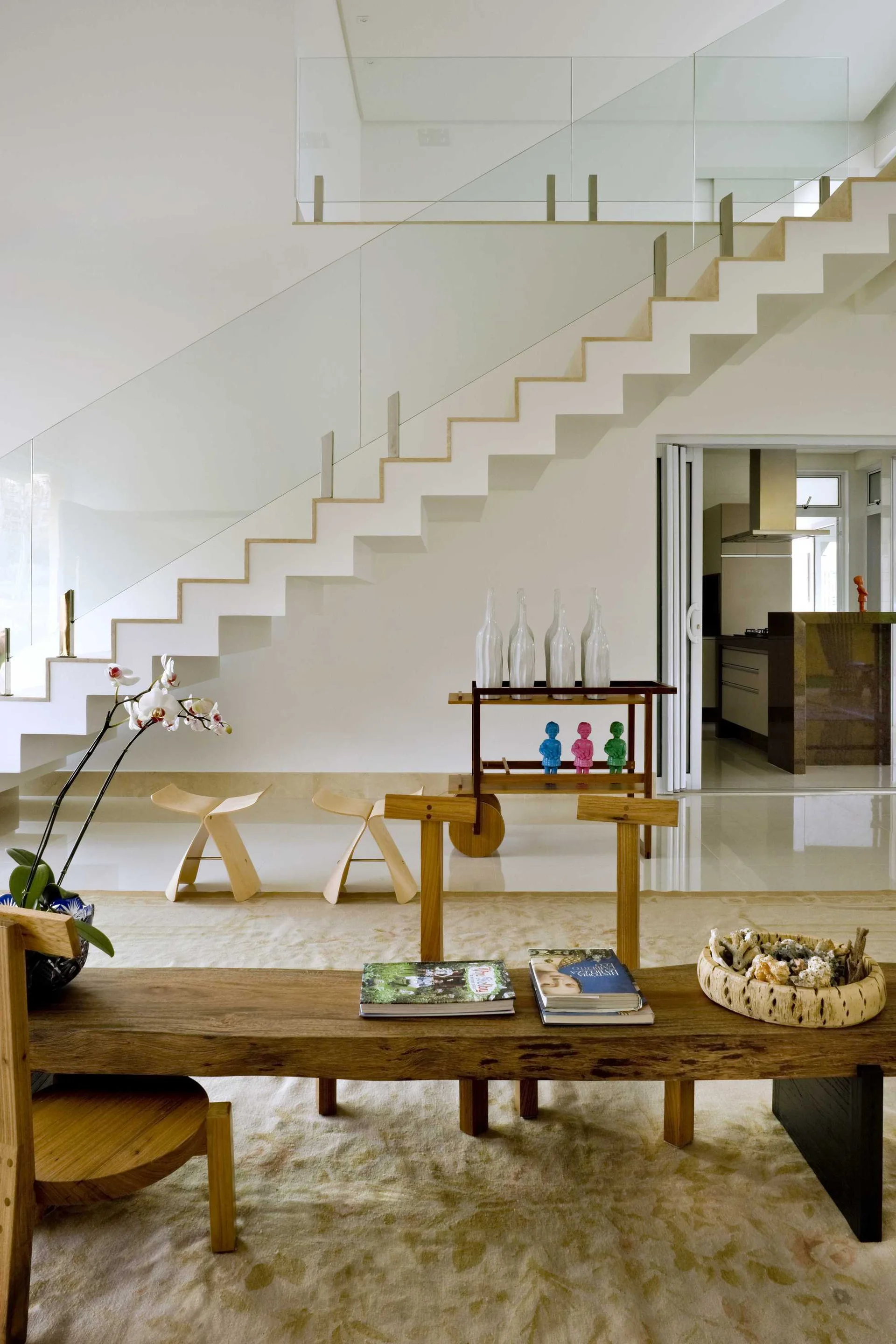 A modern living room features a white zigzag staircase with glass railings, two curved wooden stools, a rustic bench, and a polished floor.
