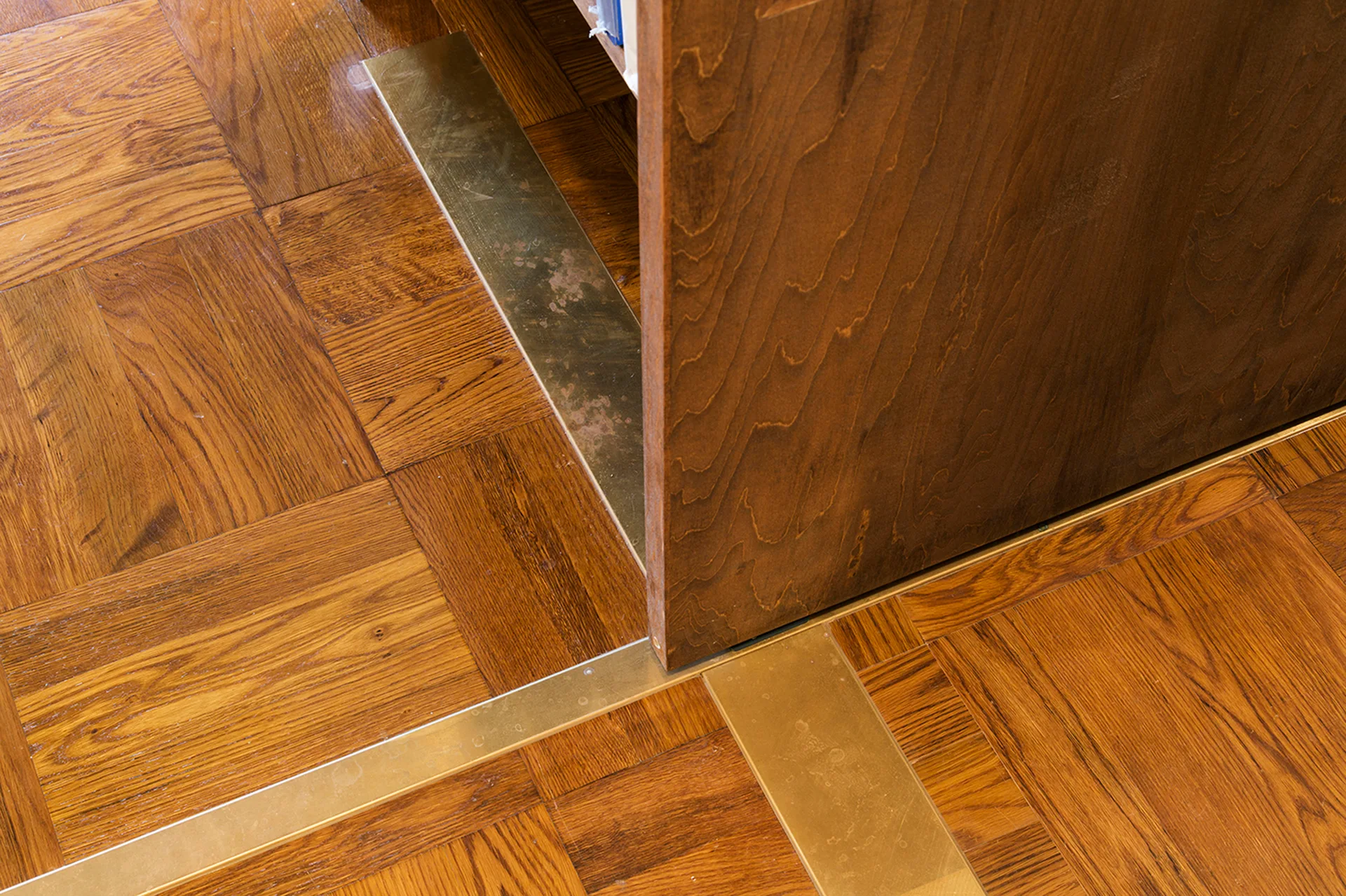 A close-up of warm brown parquet wood flooring meeting a matching wood cabinet base, separated by sleek brass metal inlay strips.