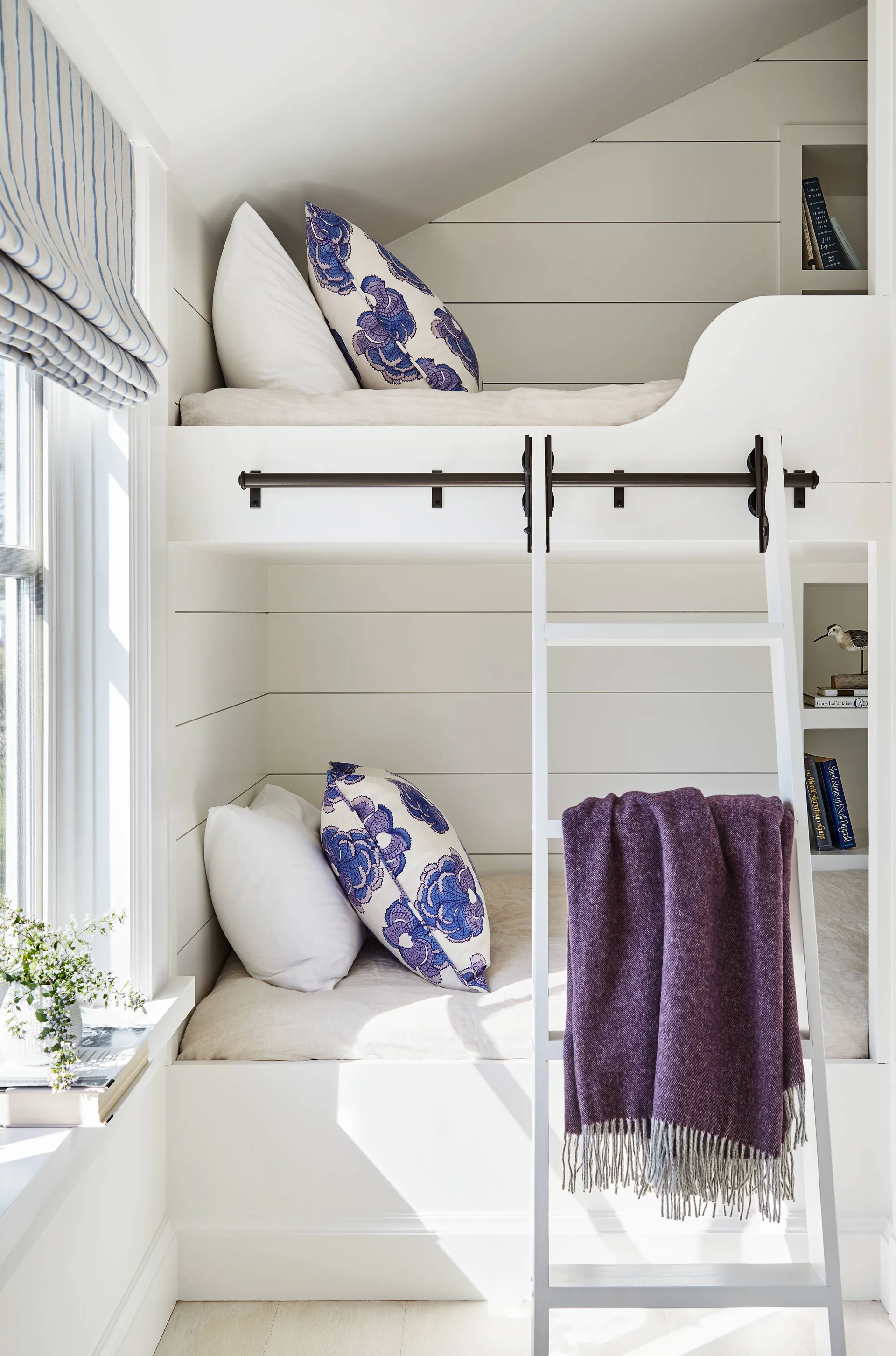 Built-in white bunk beds feature shiplap walls, integrated bookshelves, and a ladder draped with a purple throw blanket.