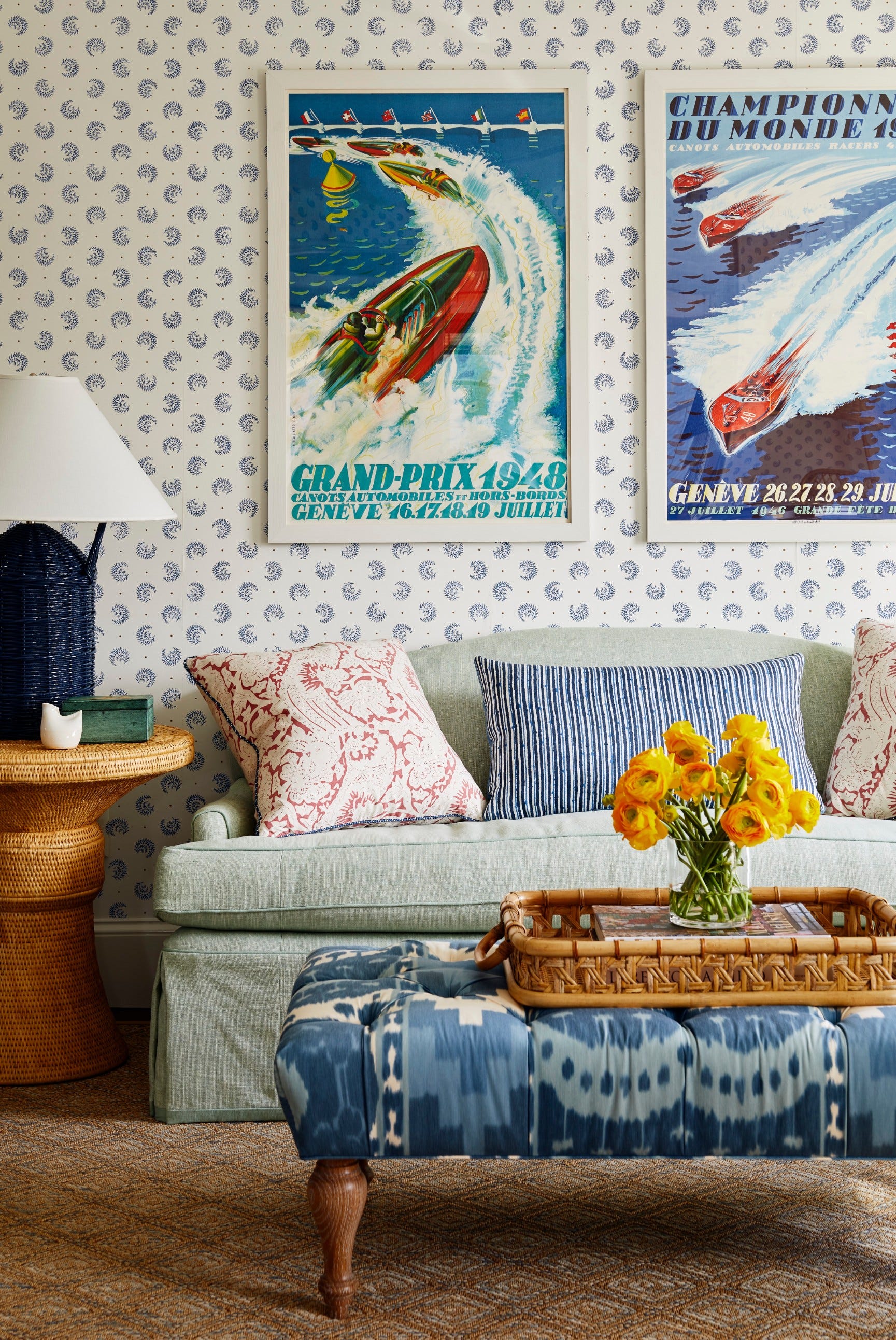 A light blue sofa with patterned pillows sits in front of patterned wallpaper and vintage posters, with a tufted ikat ottoman in the foreground.