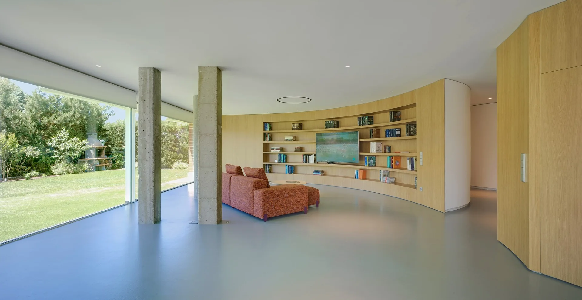 A modern living room features a curved wood paneled wall with built-in bookshelves, an orange sofa, concrete pillars, and glass walls facing a garden.