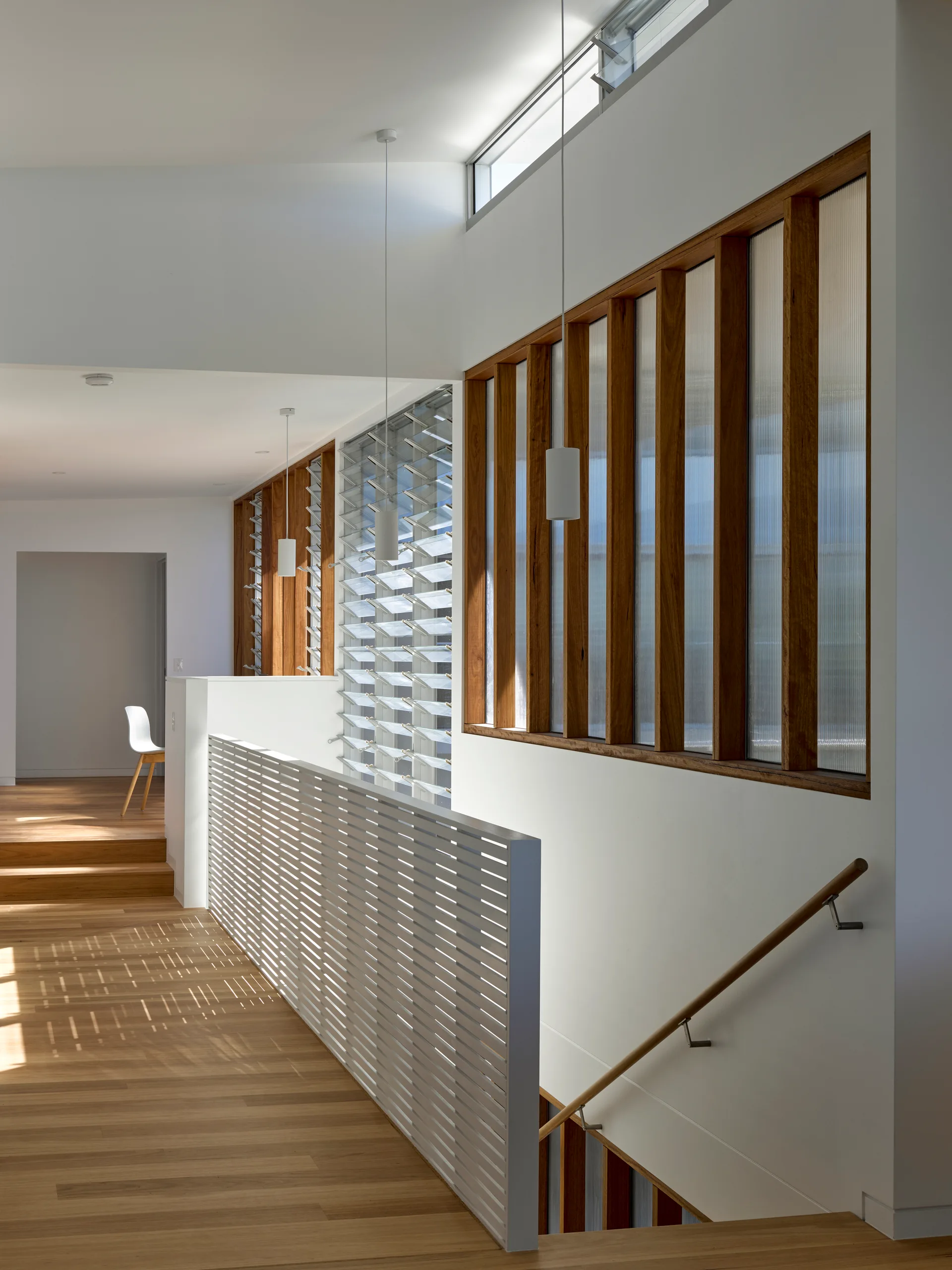 A modern stairwell landing features light wood floors, a white slatted safety railing, and tall wooden-framed windows.
