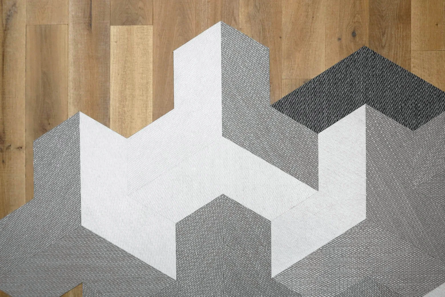An overhead close-up shows a grey and white geometric patterned rug with an irregular edge on a light brown wooden plank floor.