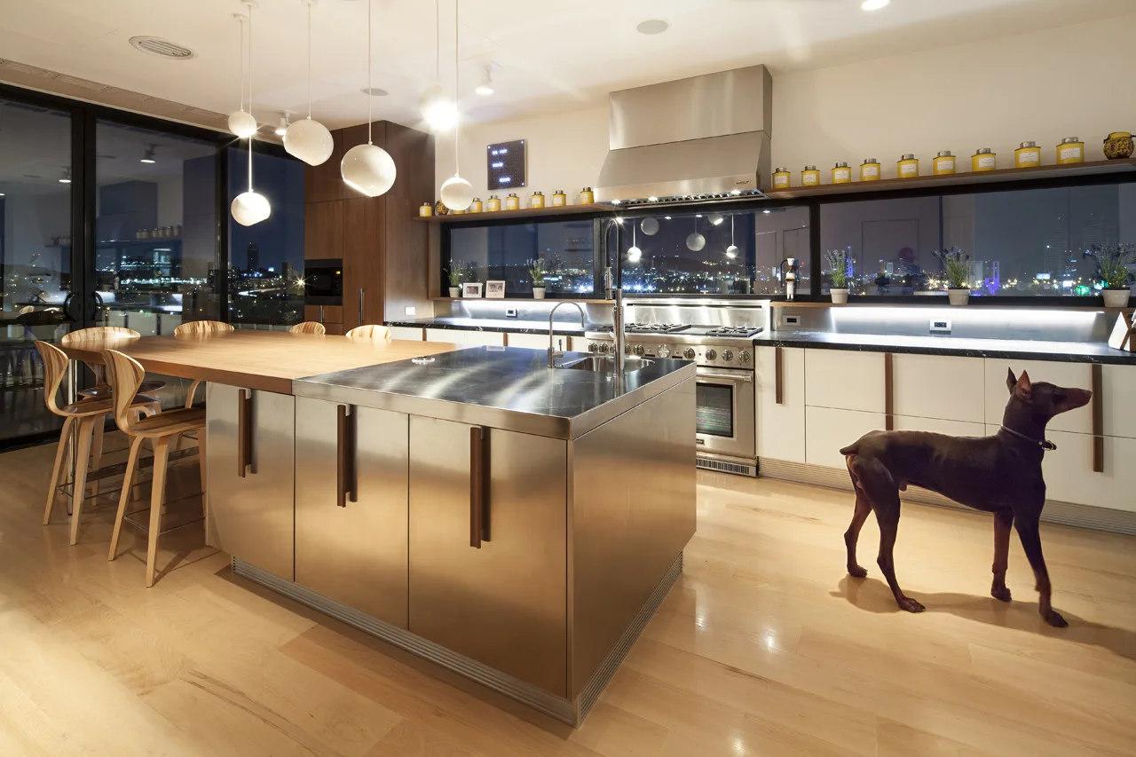 A modern kitchen features a stainless steel island with an integrated wood dining table, globe pendant lights, and a window with a nighttime city view.