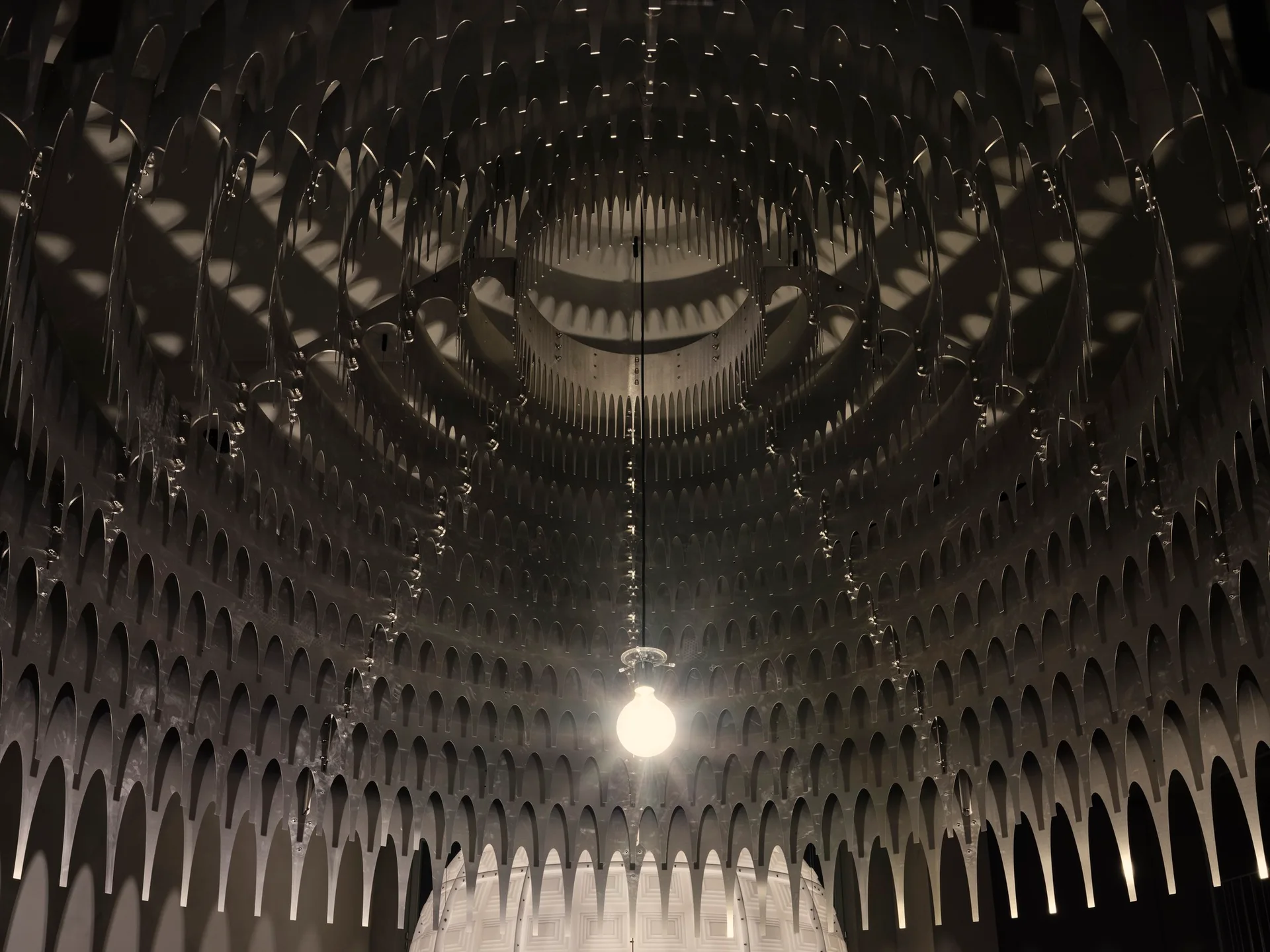 A close-up looking up at a layered grey metal ceiling installation with repeating arch cutouts and a single illuminated light bulb hanging in the center.