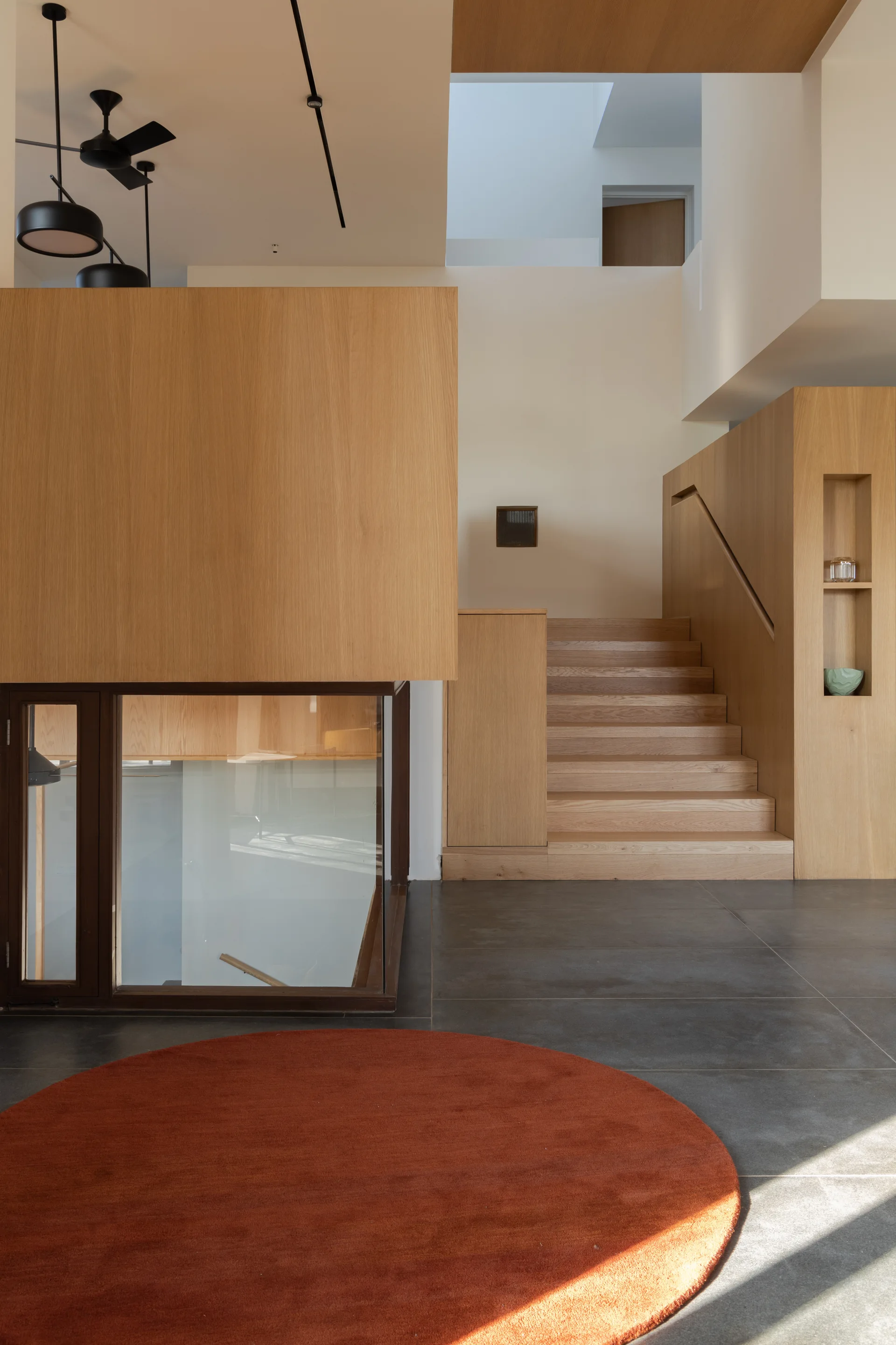 A modern light wood staircase with recessed display niches stands on dark grey floor tiles next to a circular orange rug under a bright skylight.