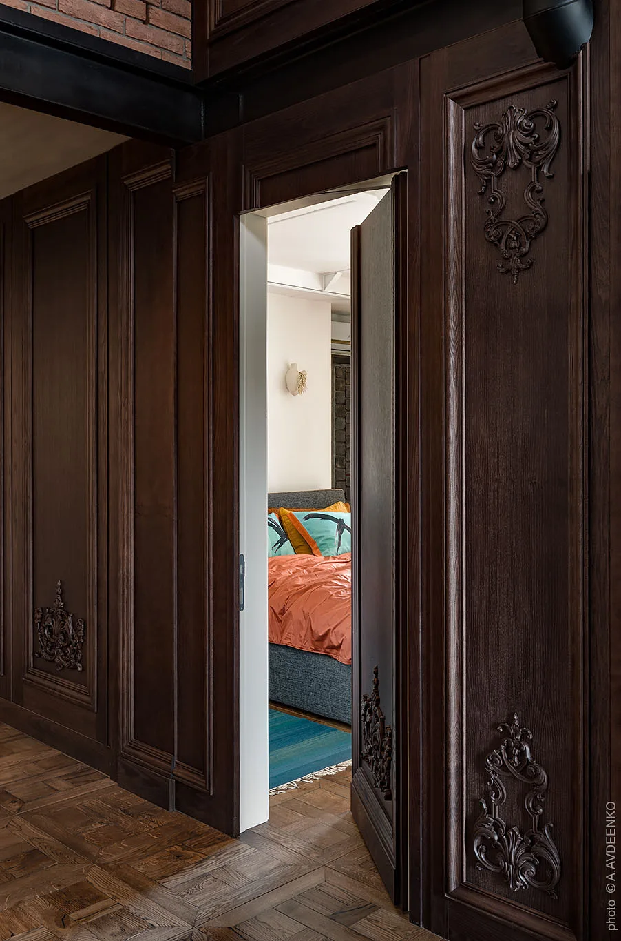 A dark wooden paneled wall with ornate carvings features an open door revealing a bright bedroom with orange bedding and a teal rug.