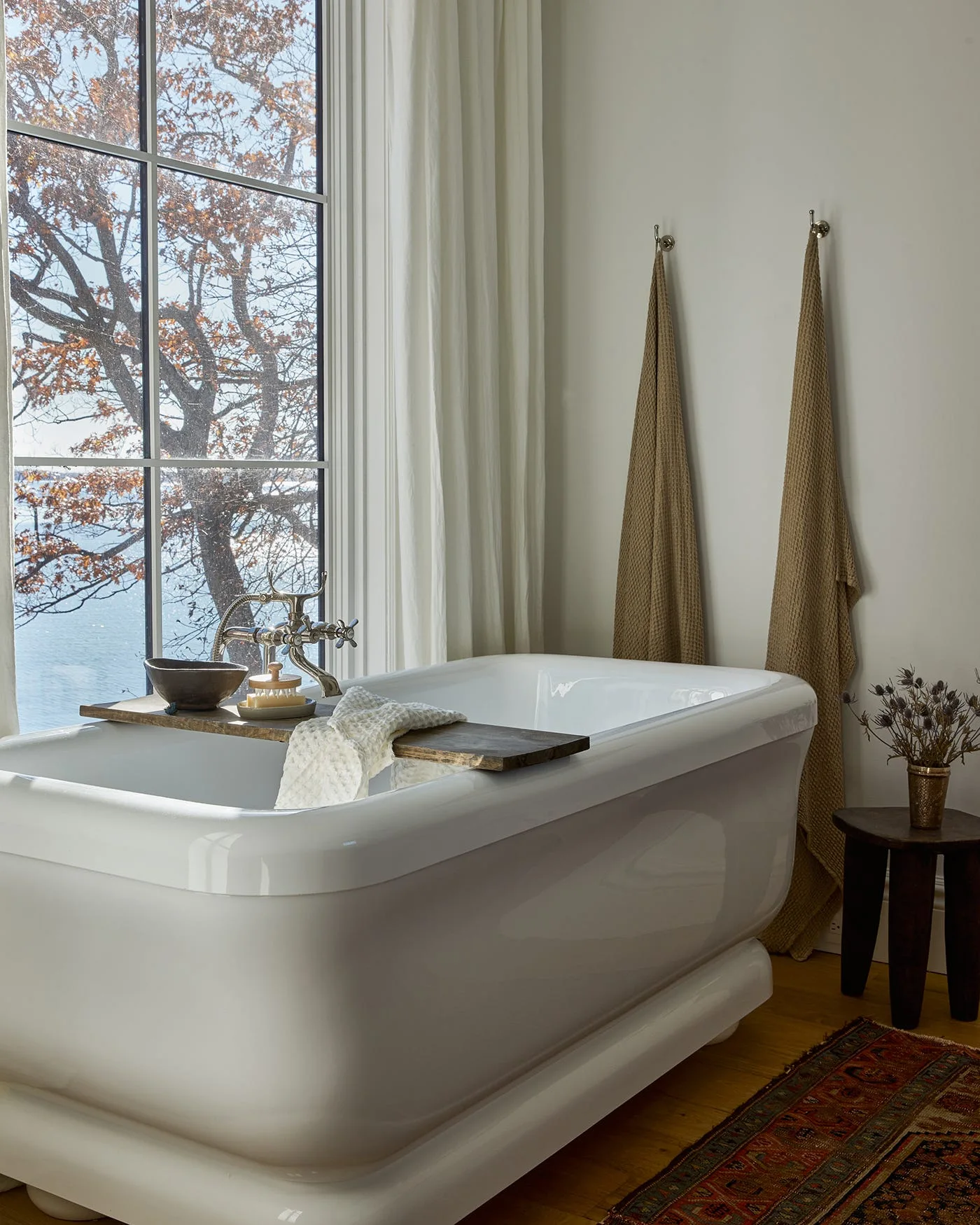 A white freestanding bathtub features a wooden tray and sits next to a large window, with two brown towels hanging on the adjacent wall.