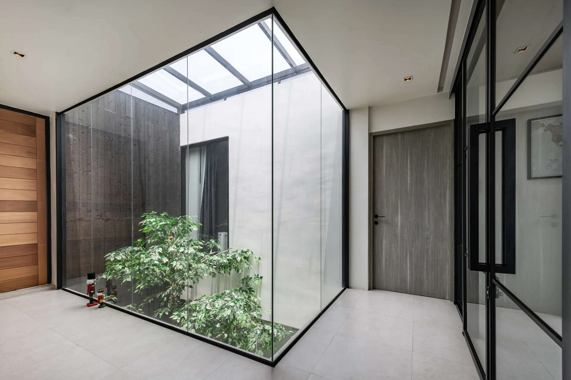 A modern hallway with light gray floor tiles features a central glass-enclosed atrium containing a small green tree beneath an overhead skylight.