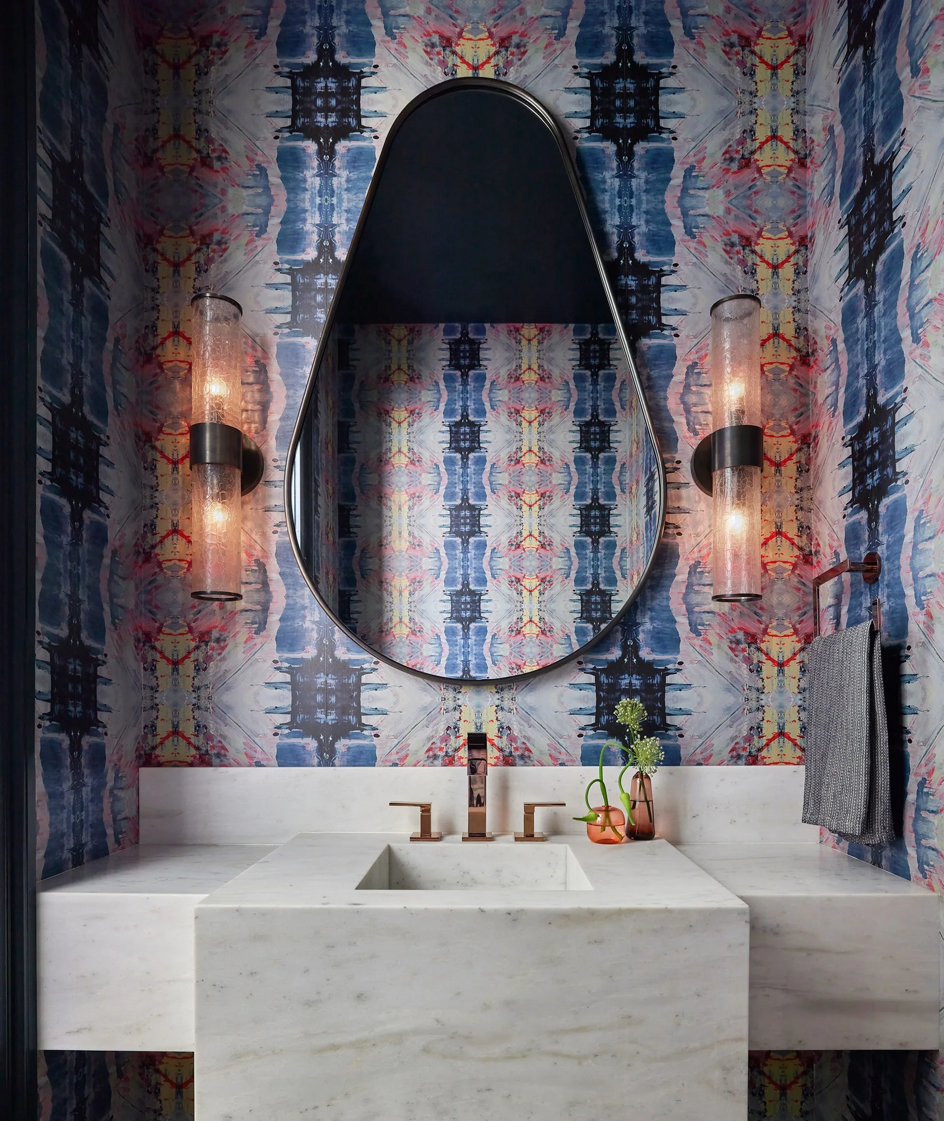 A modern powder room features a white marble sink vanity, a teardrop-shaped mirror, colorful abstract wallpaper, and two glowing glass sconces.