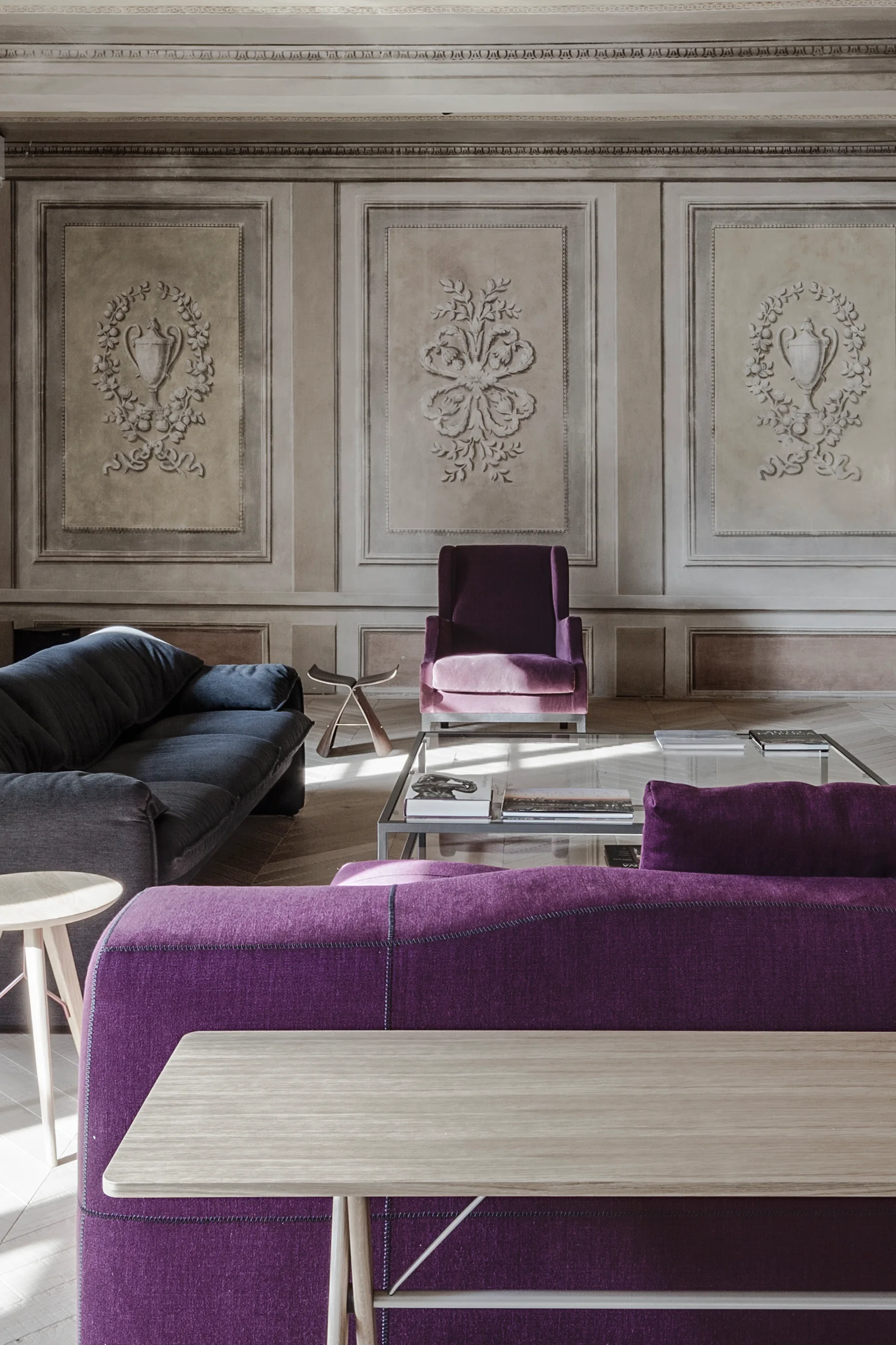 A formal living room features deep purple sofas and an armchair set against beige neoclassical paneled walls with ornate relief sculptures.