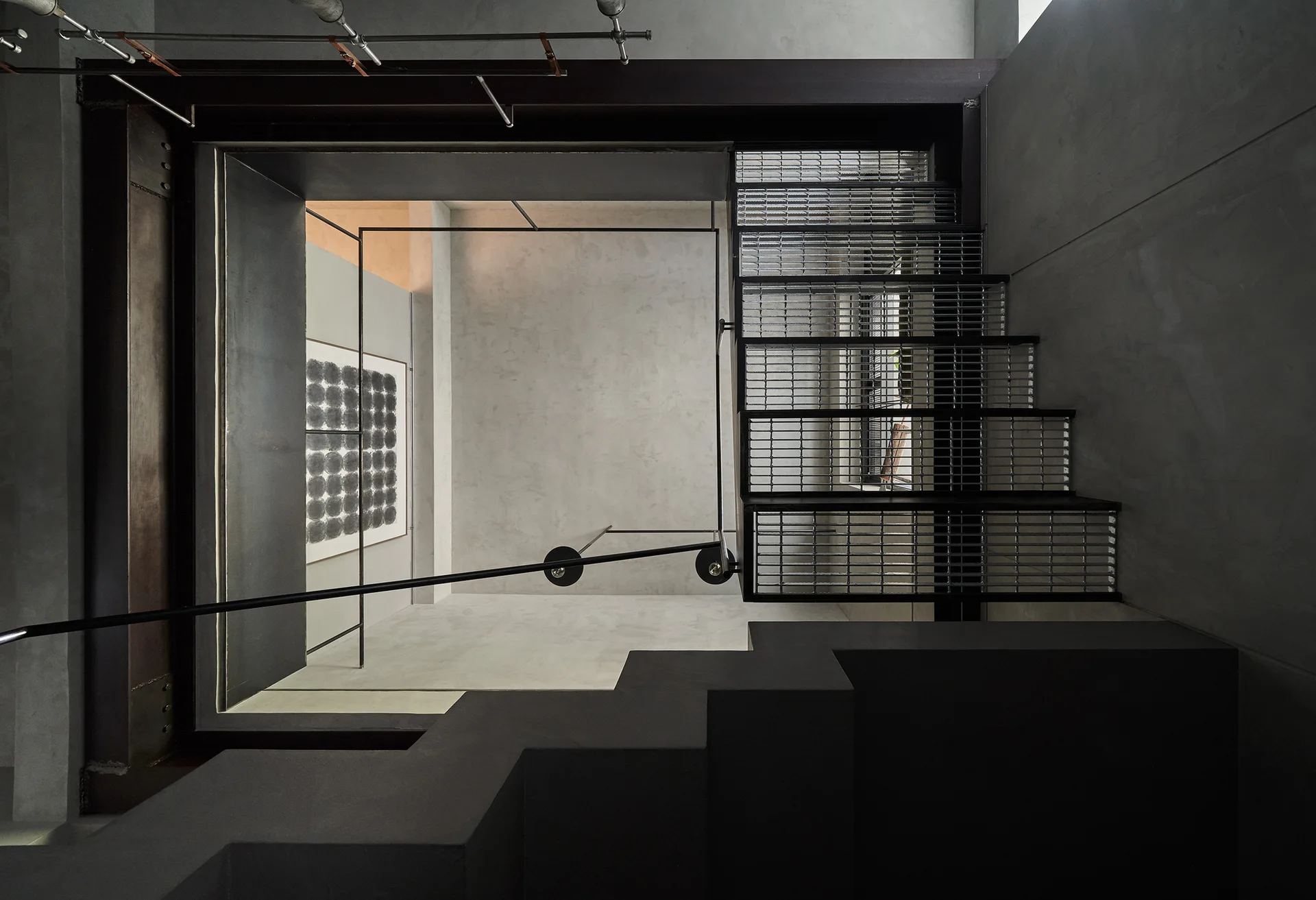 An industrial staircase with metal grating steps and a thin black railing set against smooth gray concrete walls and structural steel beams.
