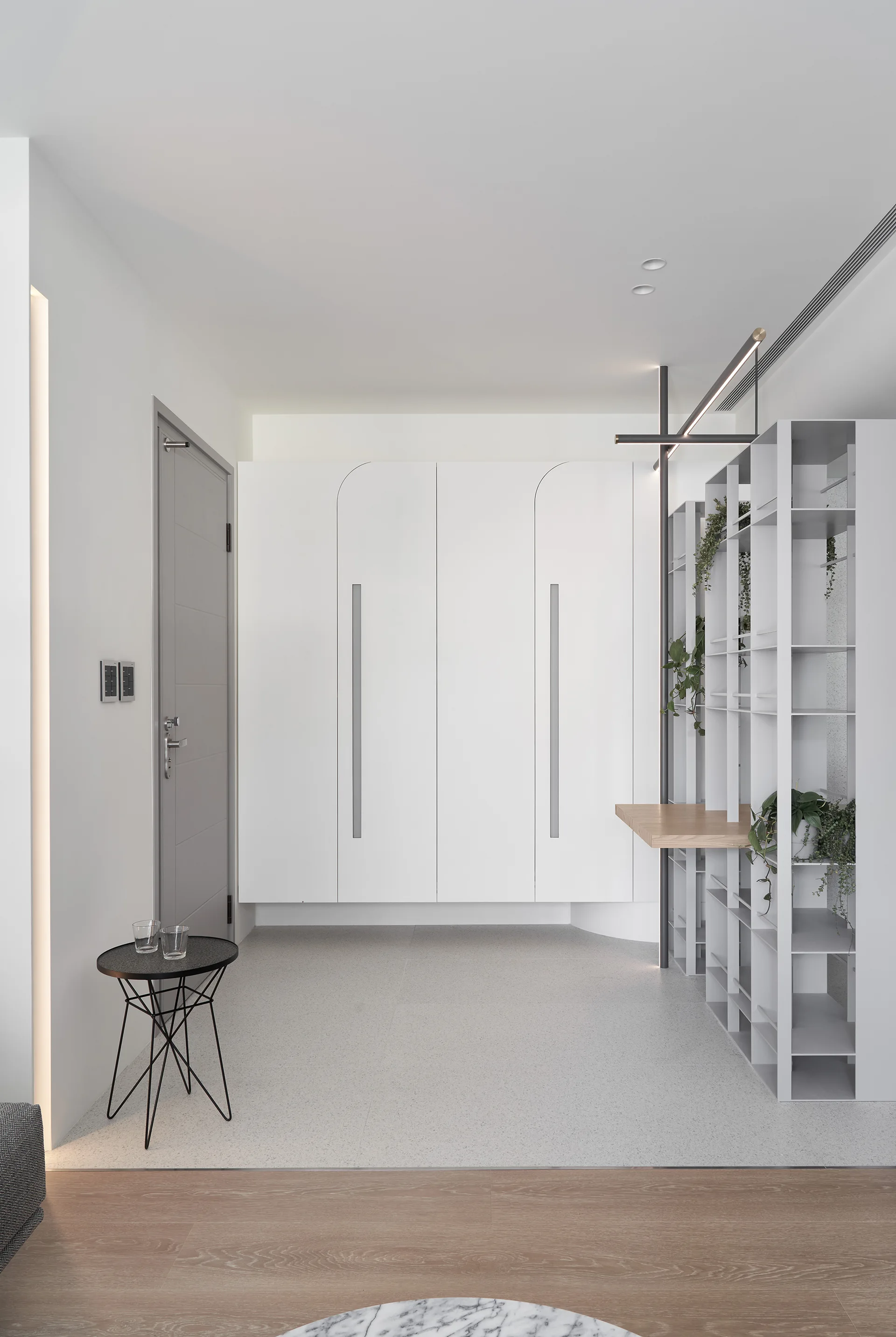 A minimalist white entryway featuring floating cabinets, a tall open metal shelving unit with plants, a grey door, and a small black side table.