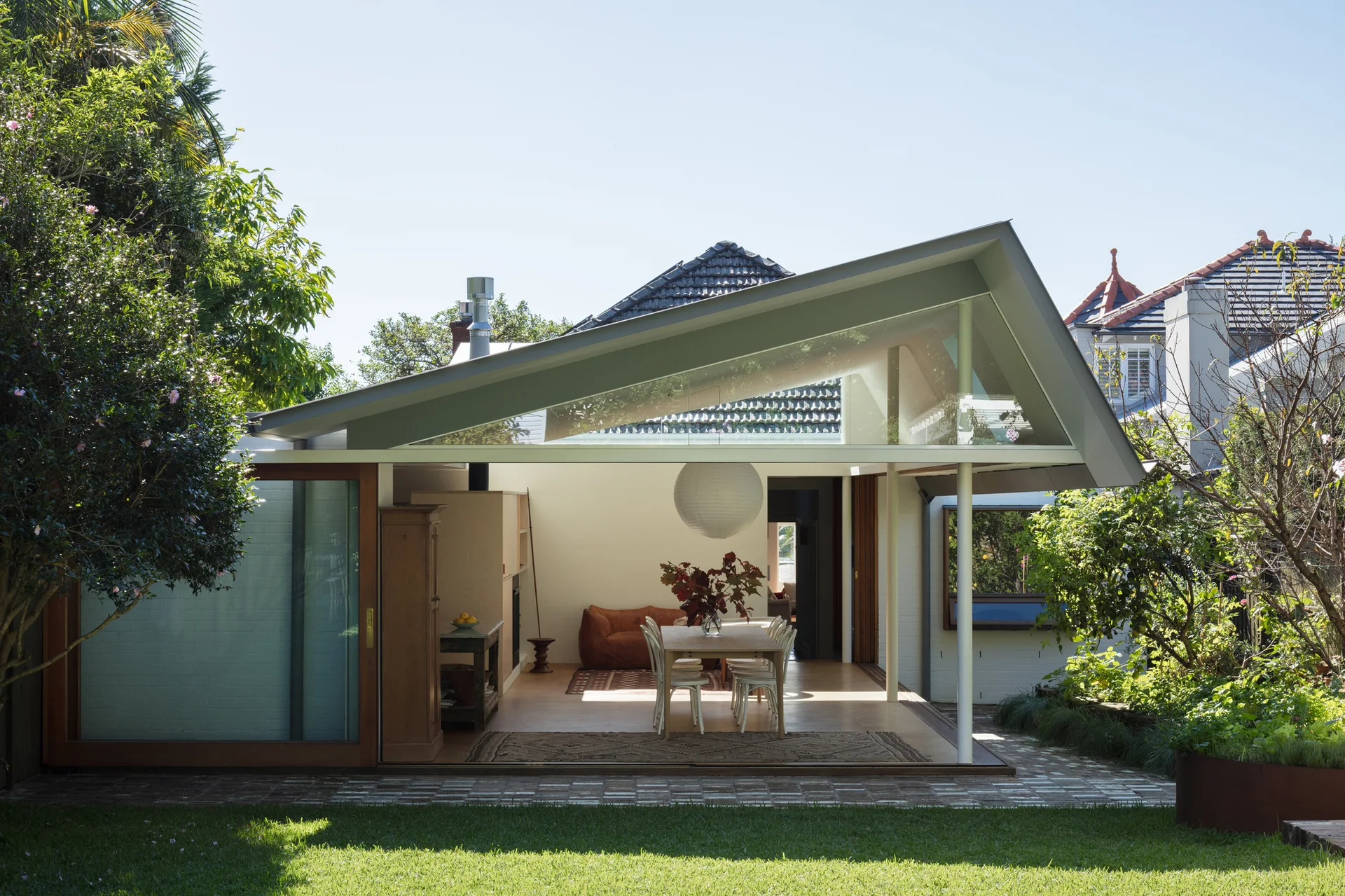 An indoor-outdoor dining room with white chairs under an angled roof overhang looks out onto a green garden.