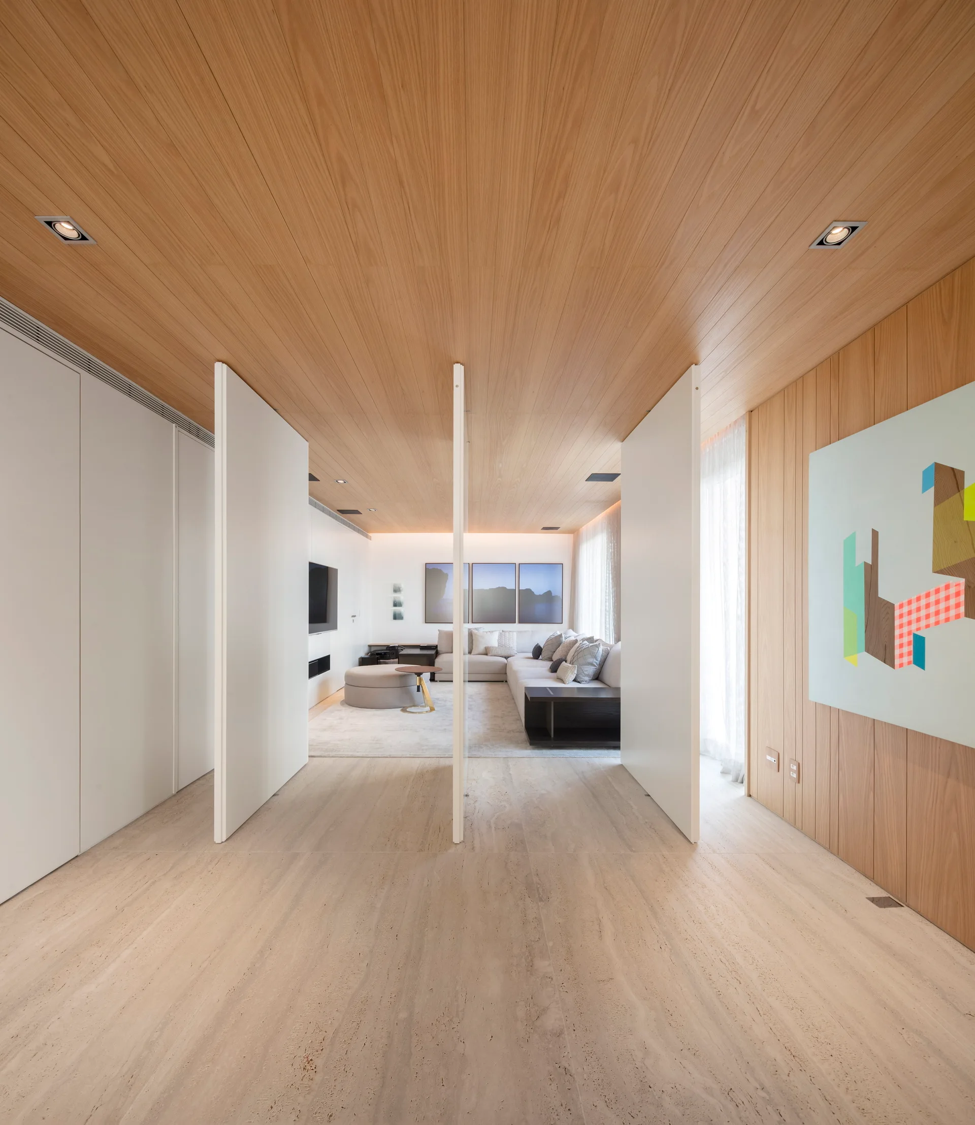 A modern interior space features light wood ceiling paneling, travertine stone flooring, and white sliding wall partitions that open to a living room.