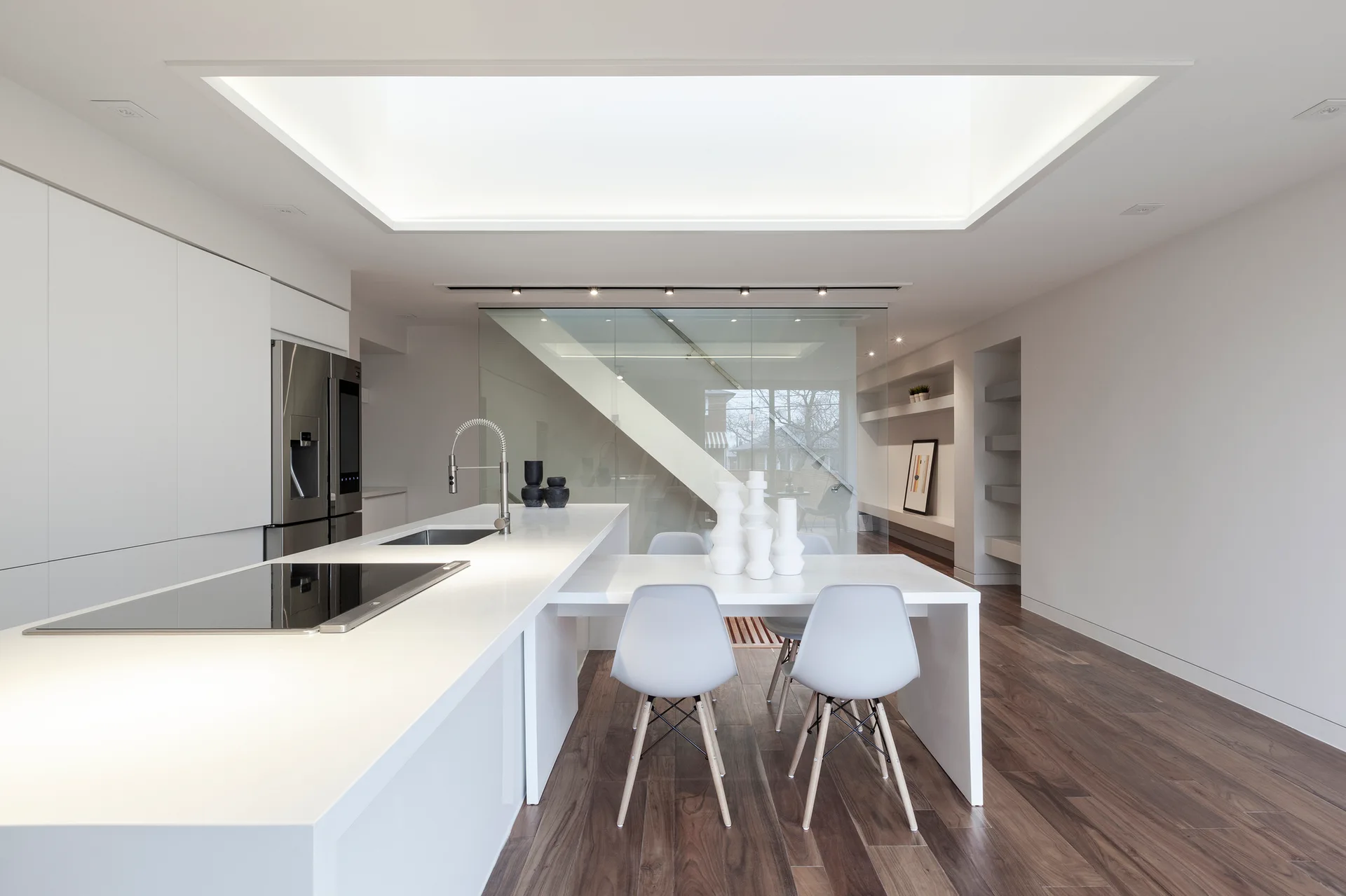 A minimalist white kitchen features an integrated dining table, a long kitchen island, and a glass partition wall over dark wood floors.
