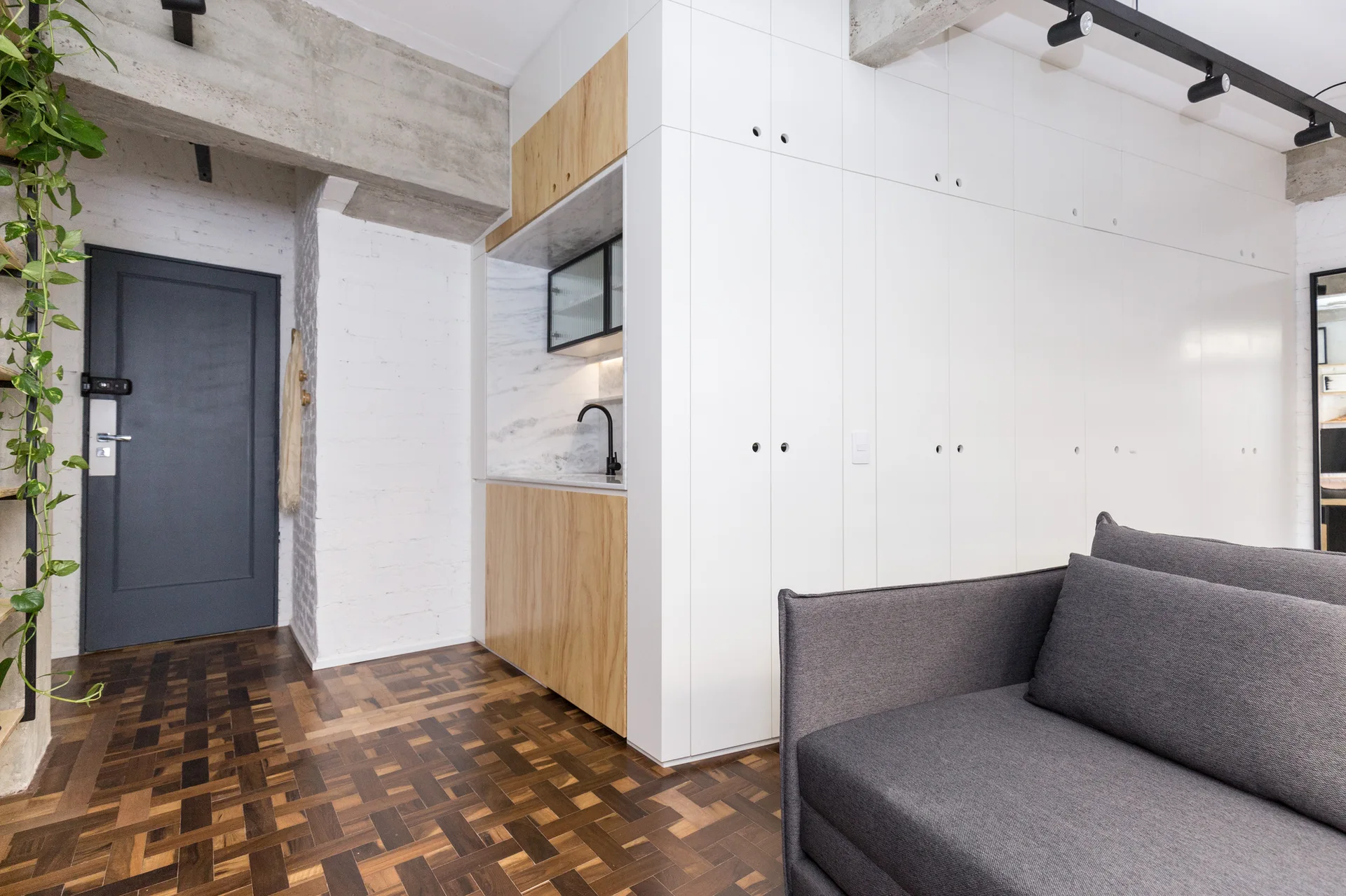 An apartment living room displays a grey sofa, parquet wood flooring, and a white built-in storage wall featuring an integrated wooden kitchenette niche.