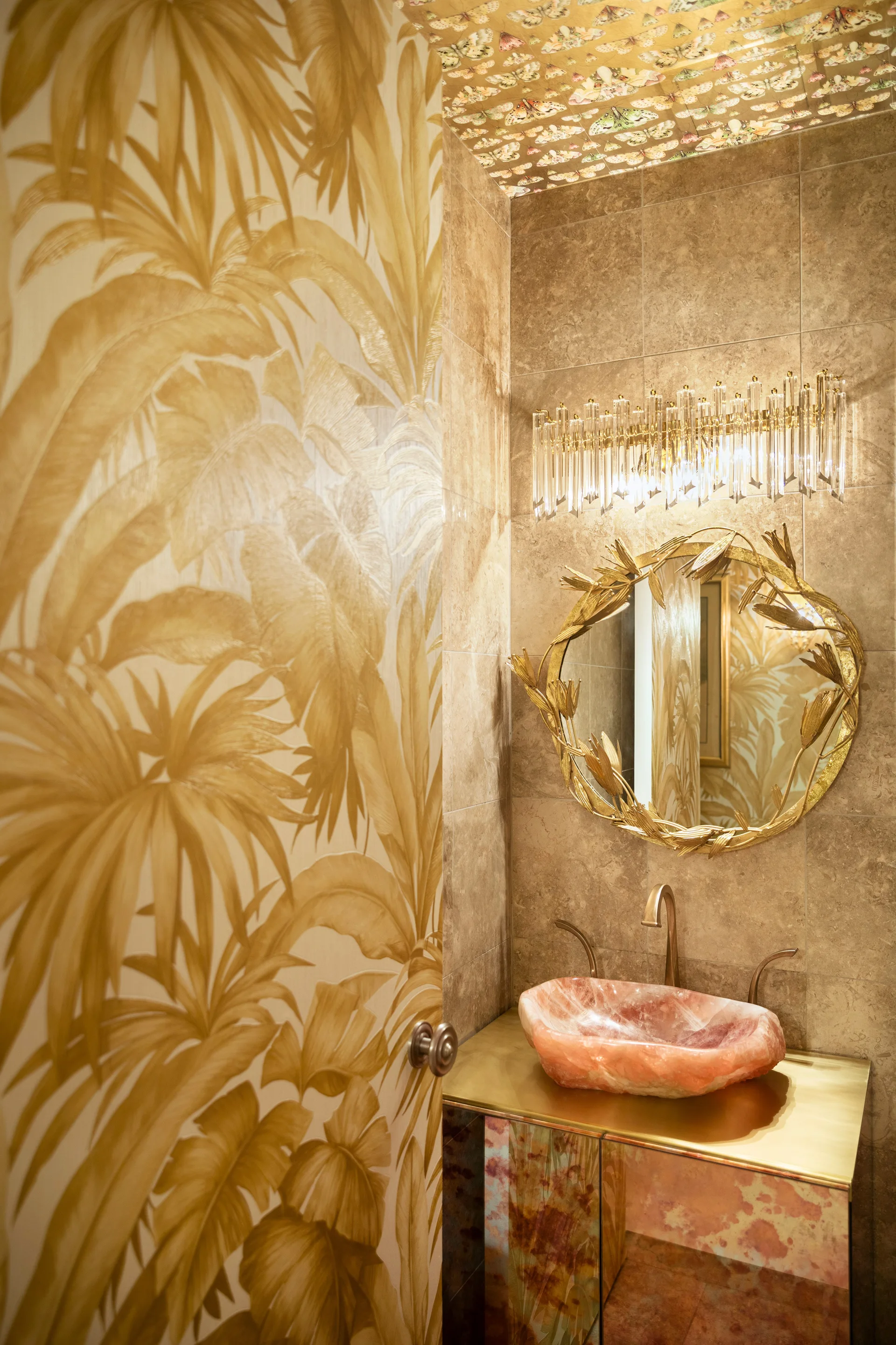 A glamorous powder room features a pink stone vessel sink, a metallic gold vanity, and botanical wallpaper with an ornate gold mirror.