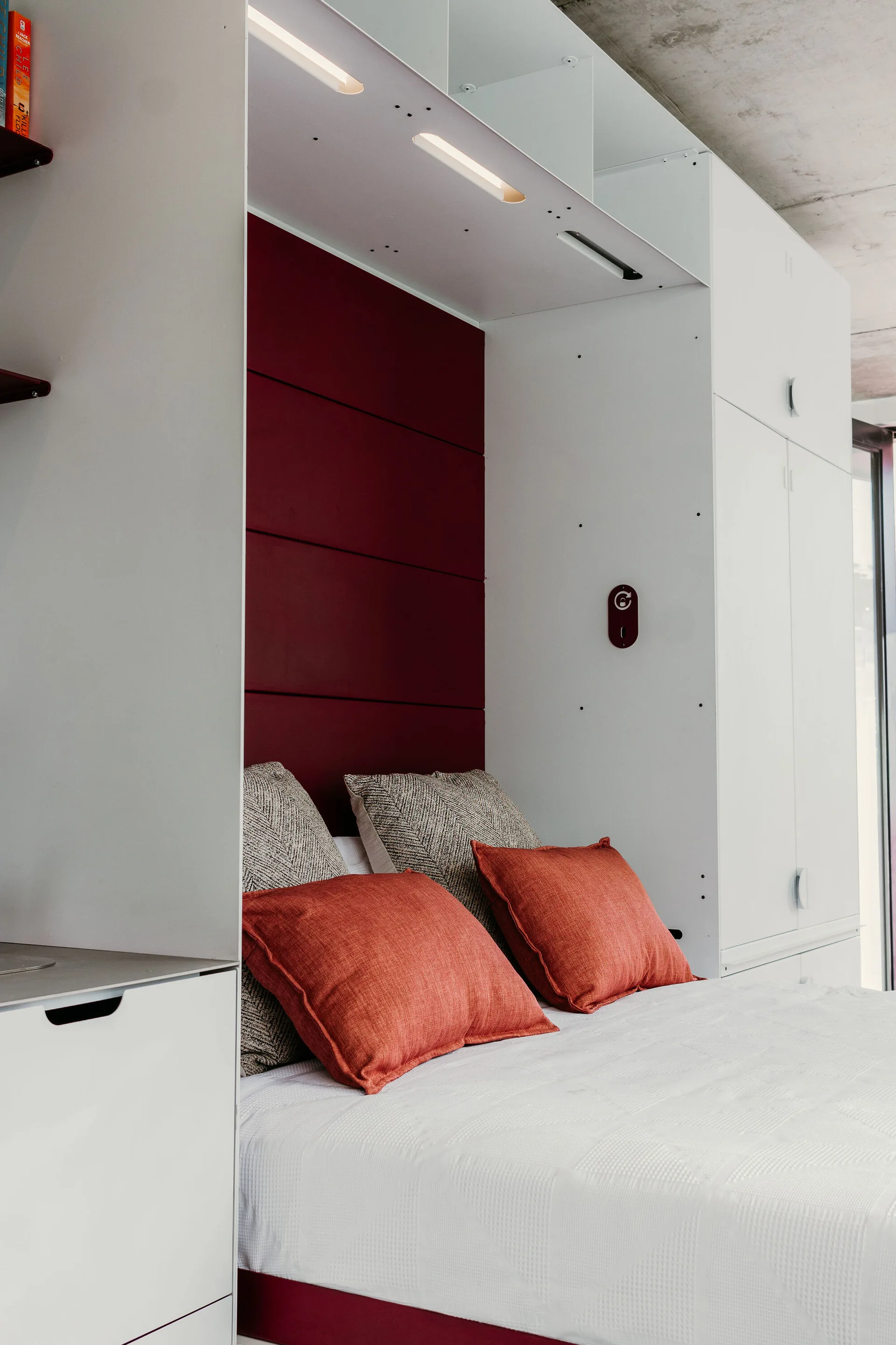 A modern bedroom features a white built-in cabinet system framing a bed with red pillows, a deep maroon headboard, and an exposed concrete ceiling.