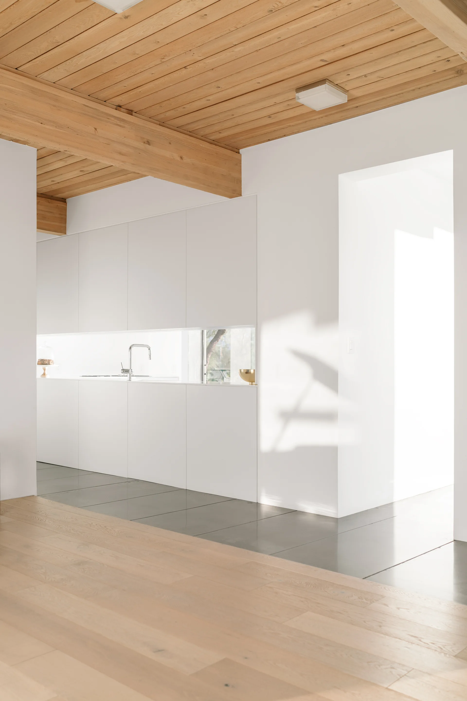 Minimalist white kitchen features flat-panel handleless cabinets, a low horizontal ribbon window backsplash, and an exposed wood plank ceiling.
