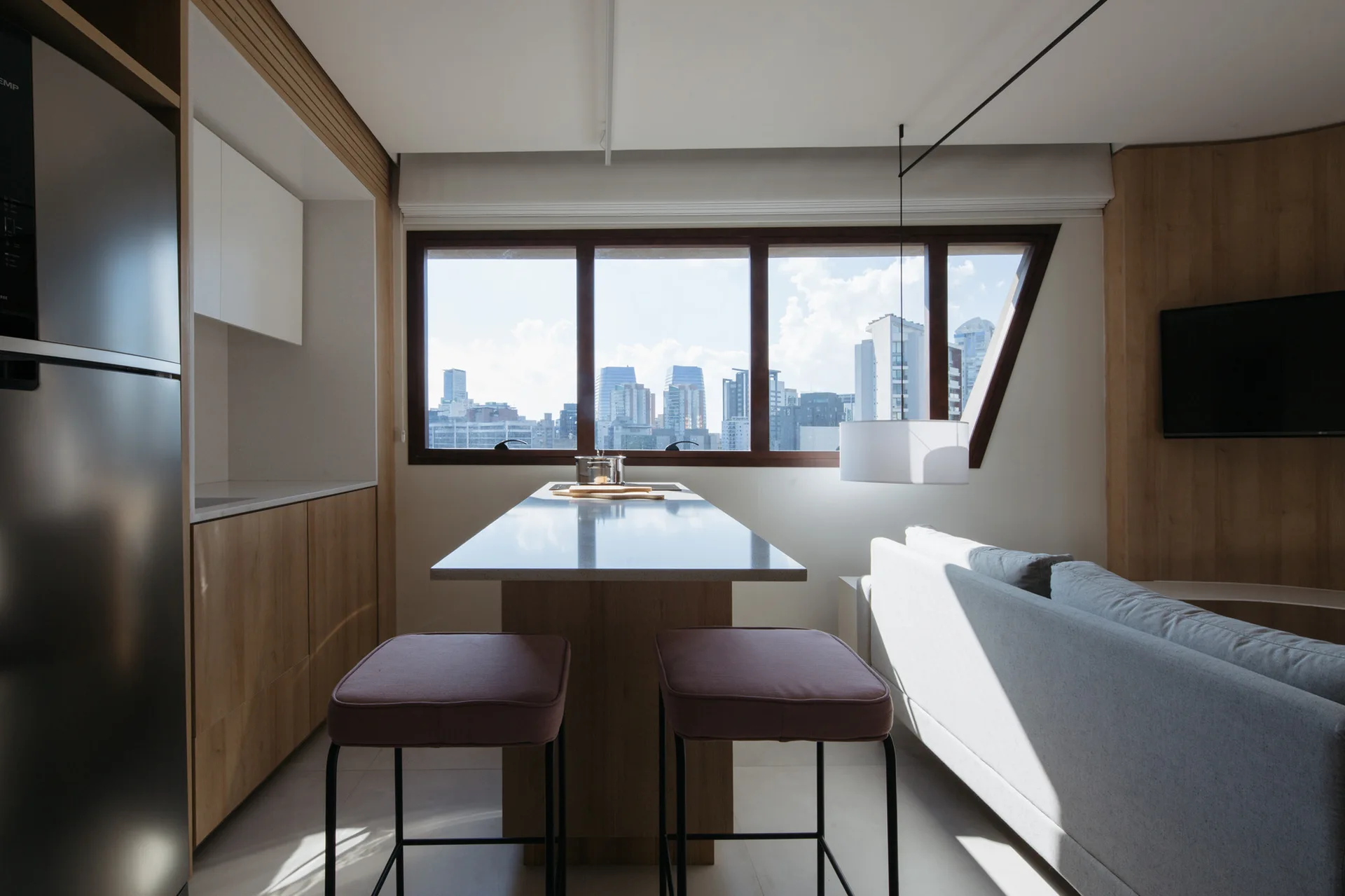 A modern apartment kitchen with a central wood island, two mauve counter stools, and a large window framing a city skyline view.