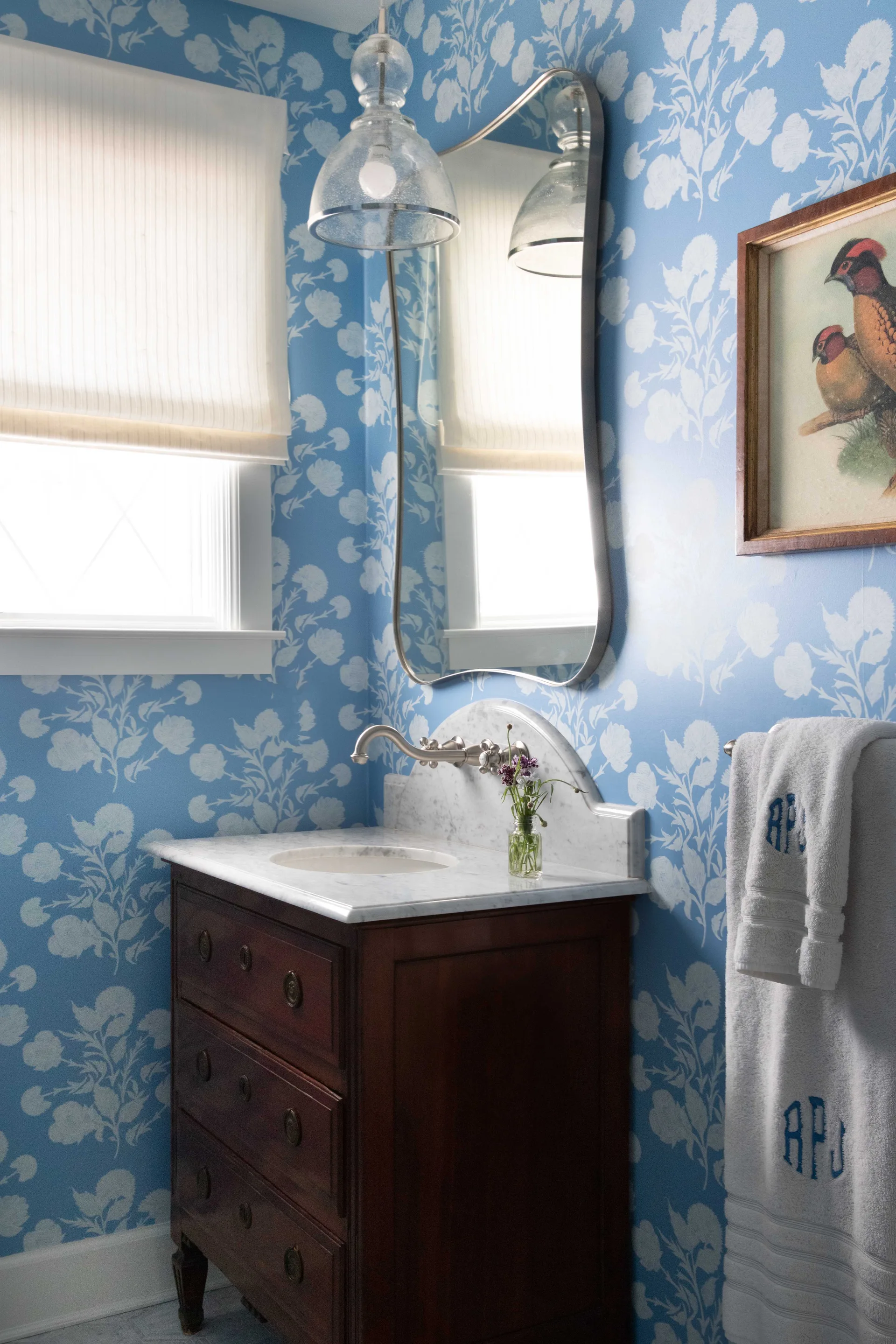 A powder room features a dark wood vanity with a marble countertop against blue floral wallpaper, a curved mirror, and a glass pendant light.