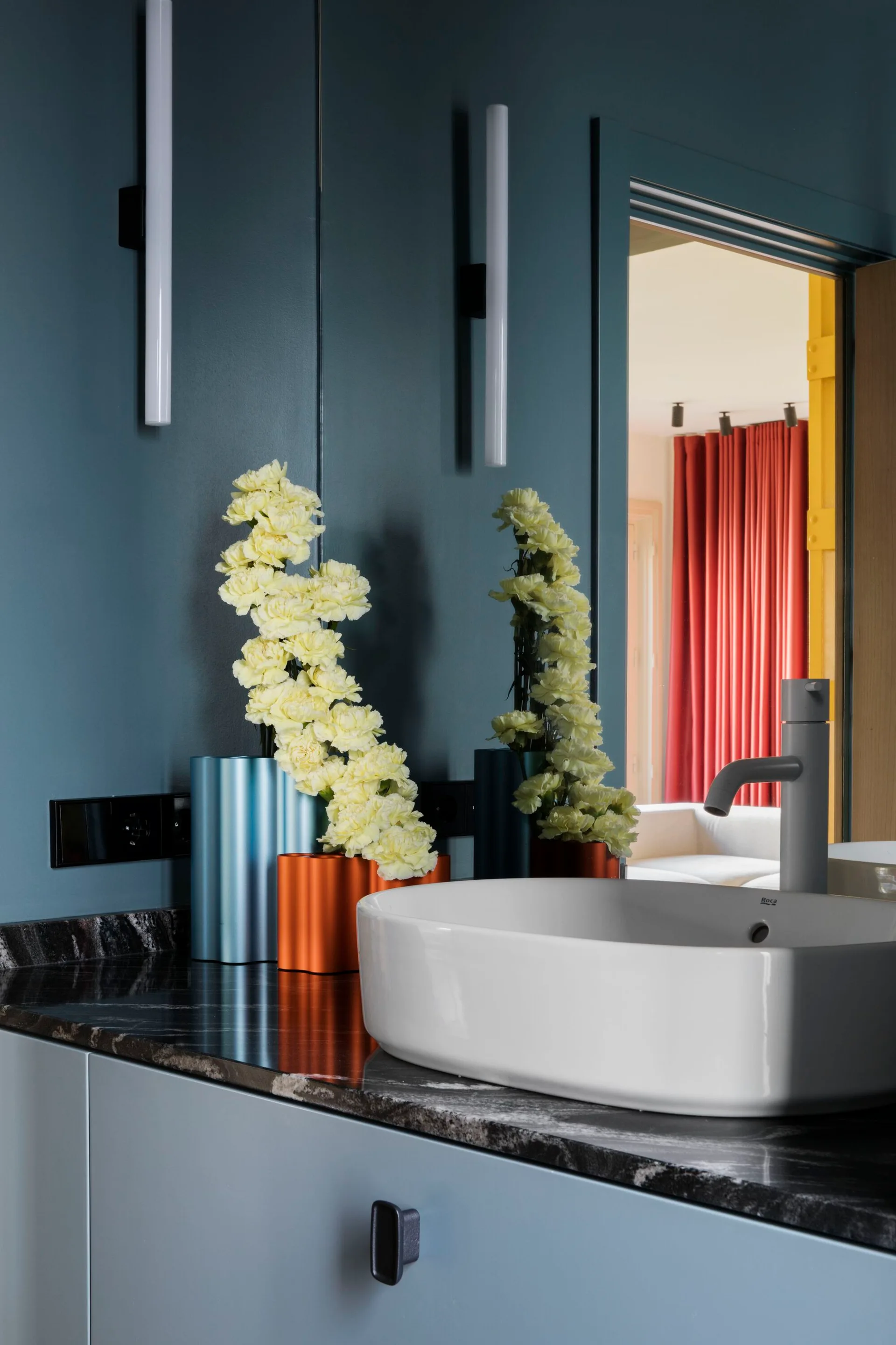 A white ceramic vessel sink sits on a dark veined marble countertop against a deep blue bathroom wall with modern vertical wall sconces.