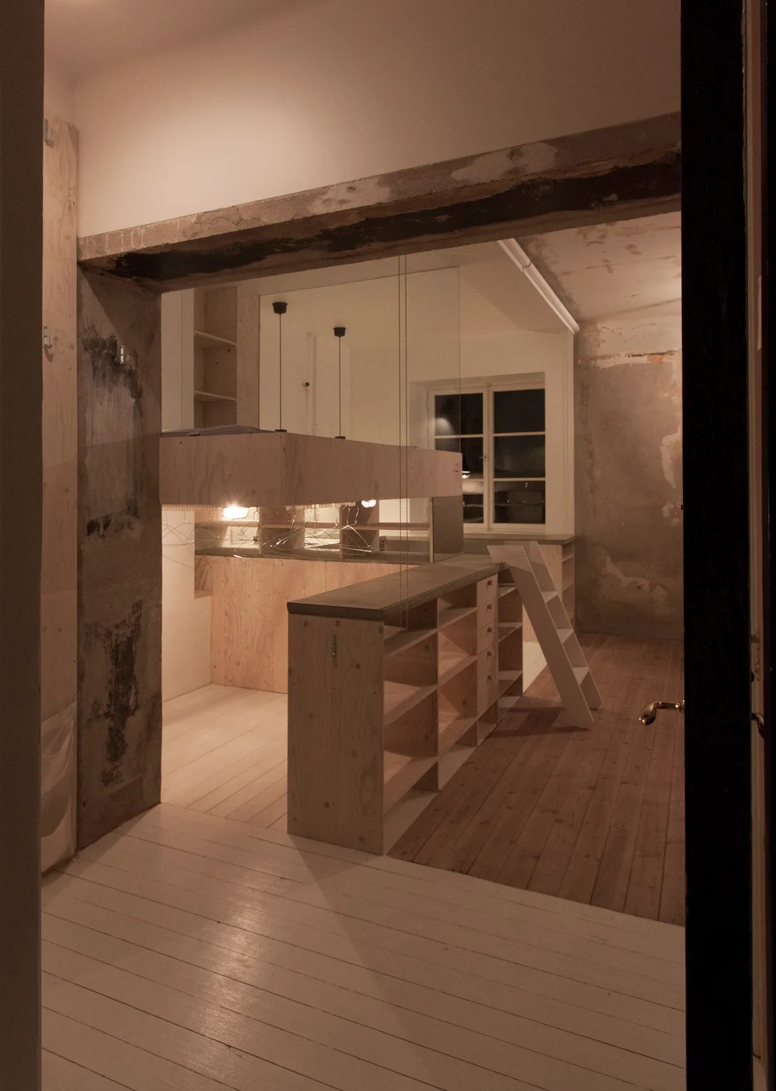 Custom plywood storage cabinets with a white ladder are shown beneath an exposed structural beam in an unfinished room with wood floors.