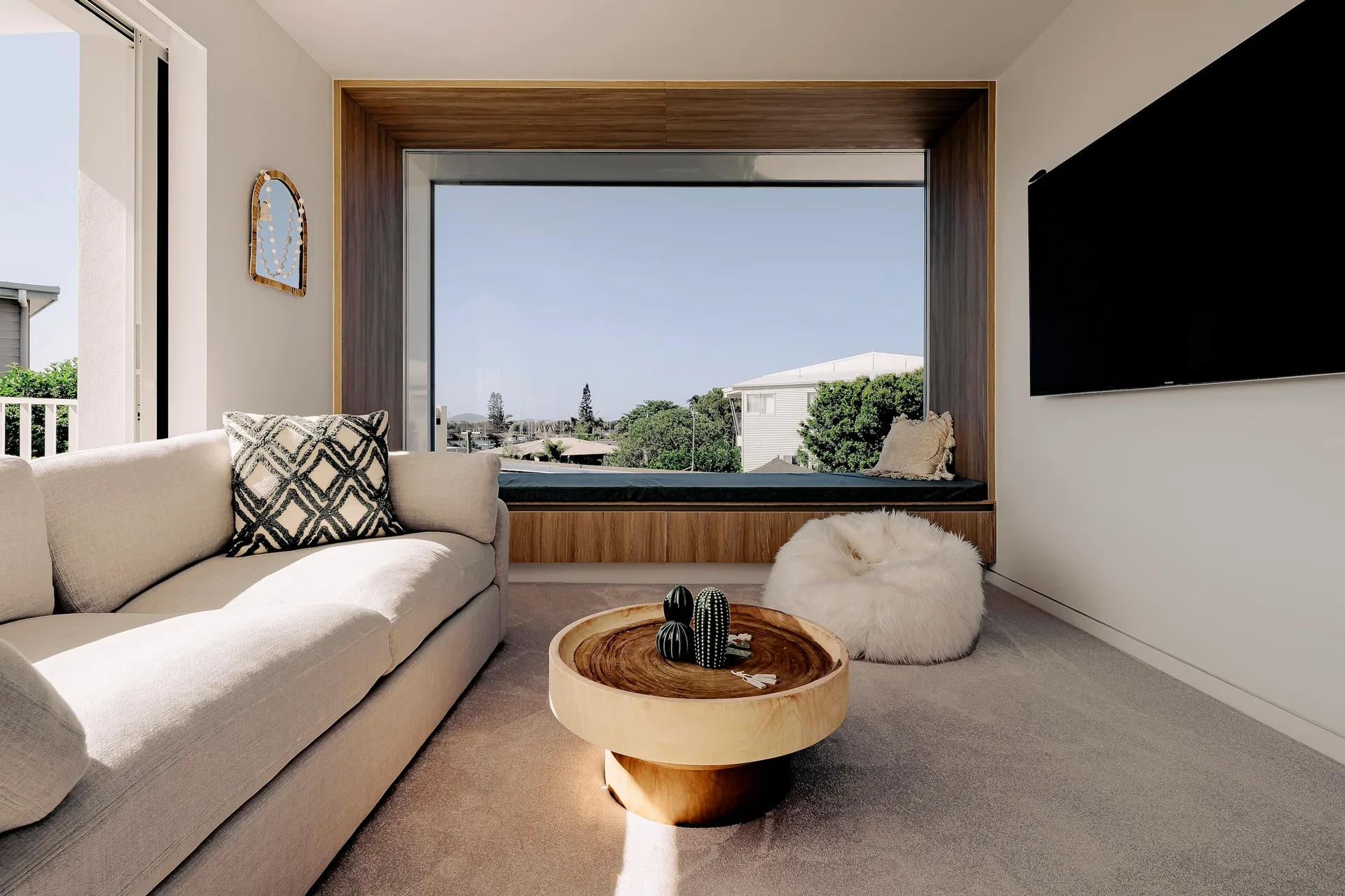 A modern living room featuring a beige sofa, a round wooden coffee table, and a large window framed in a deep wooden built-in seat.